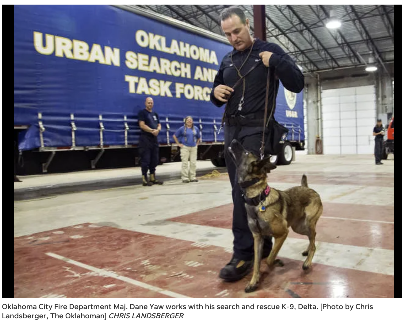 Oklahoma City Fire Department Maj. Dane Yaw with his search and rescue K-9, Delta, inside a large facility. Delta is sitting on the ground and biting Dane Yaw's pant leg, while Dane is looking down at him. Several people are standing in the background near a large truck with a blue cover that reads "Oklahoma Urban Search and Task Force." The setting appears to be a spacious indoor training or operational area.