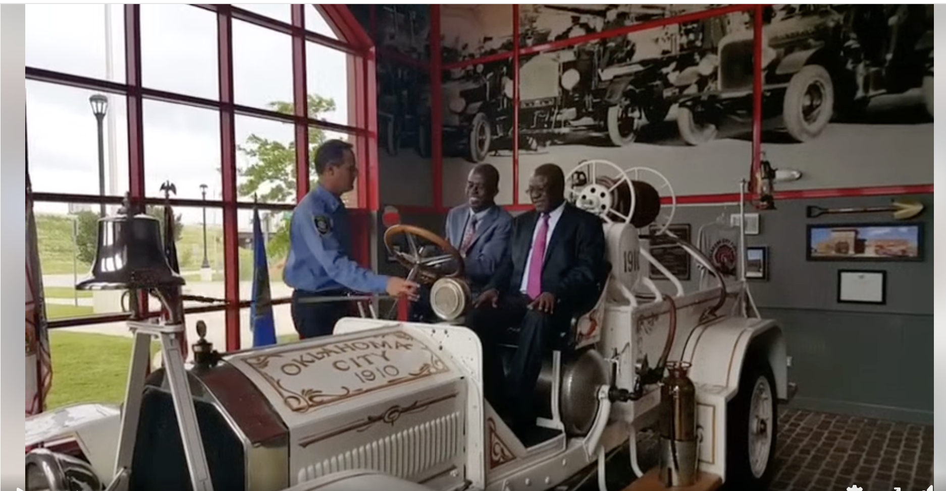 Three men, one in a police uniform and two in suits, are sitting in a vintage fire truck exhibit at a museum, with a large black-and-white photo of fire trucks and framed pictures on the wall behind them.
