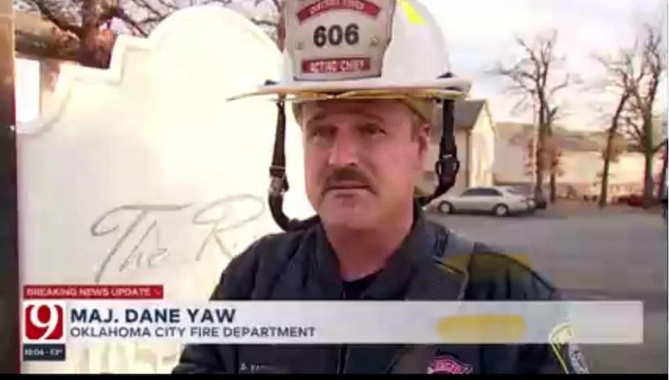 A firefighter, MAJ. DANE YAW from the Oklahoma City Fire Department, wearing a helmet and uniform, providing a news interview outdoors with trees and buildings in the background.