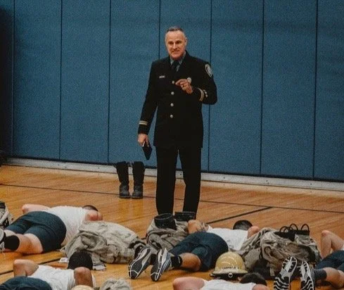 A police officer standing on a gymnasium floor speaking to children lying on the floor, with some children in military-style uniforms and helmets.