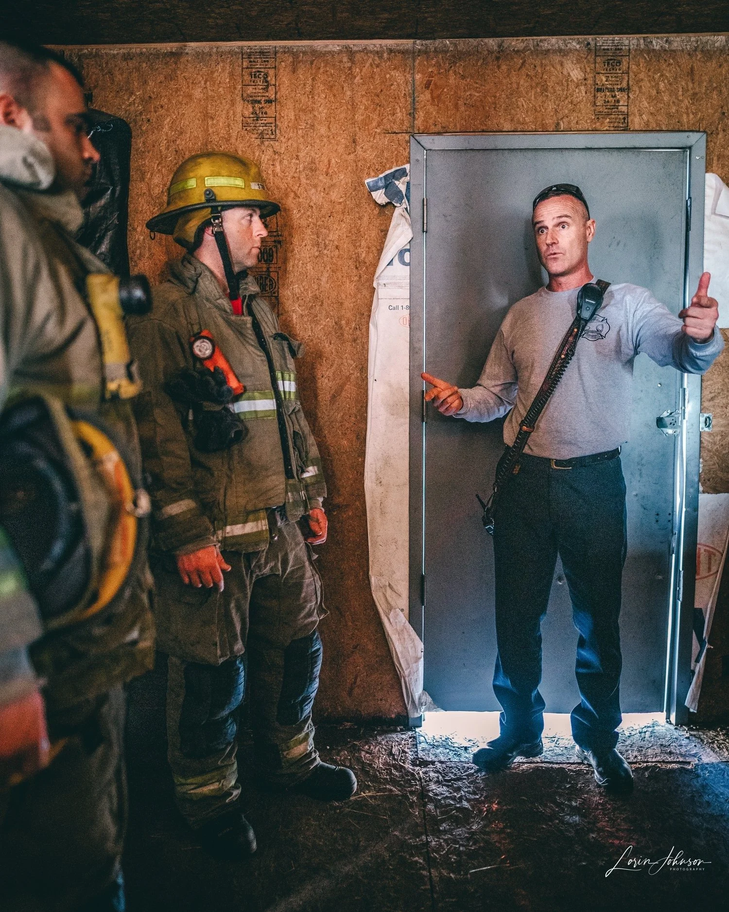 A firefighter giving instructions to a group of firefighters inside a fire station or training facility, with a door in the background.