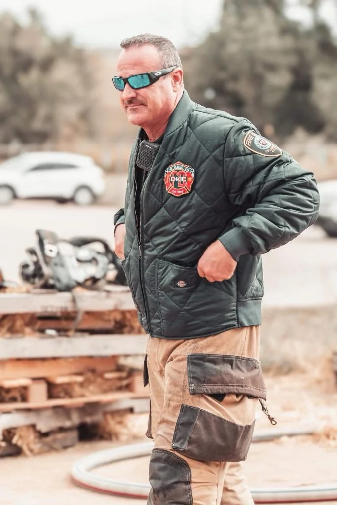 A man wearing sunglasses, a black quilted jacket with fire department patches, and cargo pants, standing outdoors near a pile of wood and some parked cars.