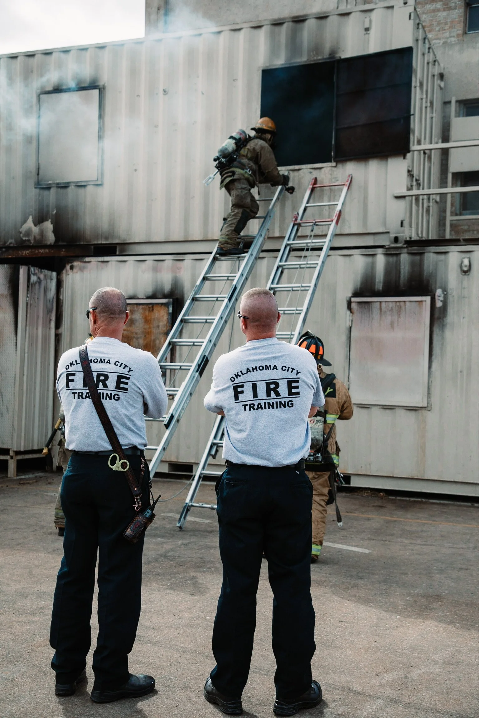 Firefighter training exercise with firemen observing a firefighter practicing on a ladder at a mock fire scene, with a burning building made of shipping containers in the background.