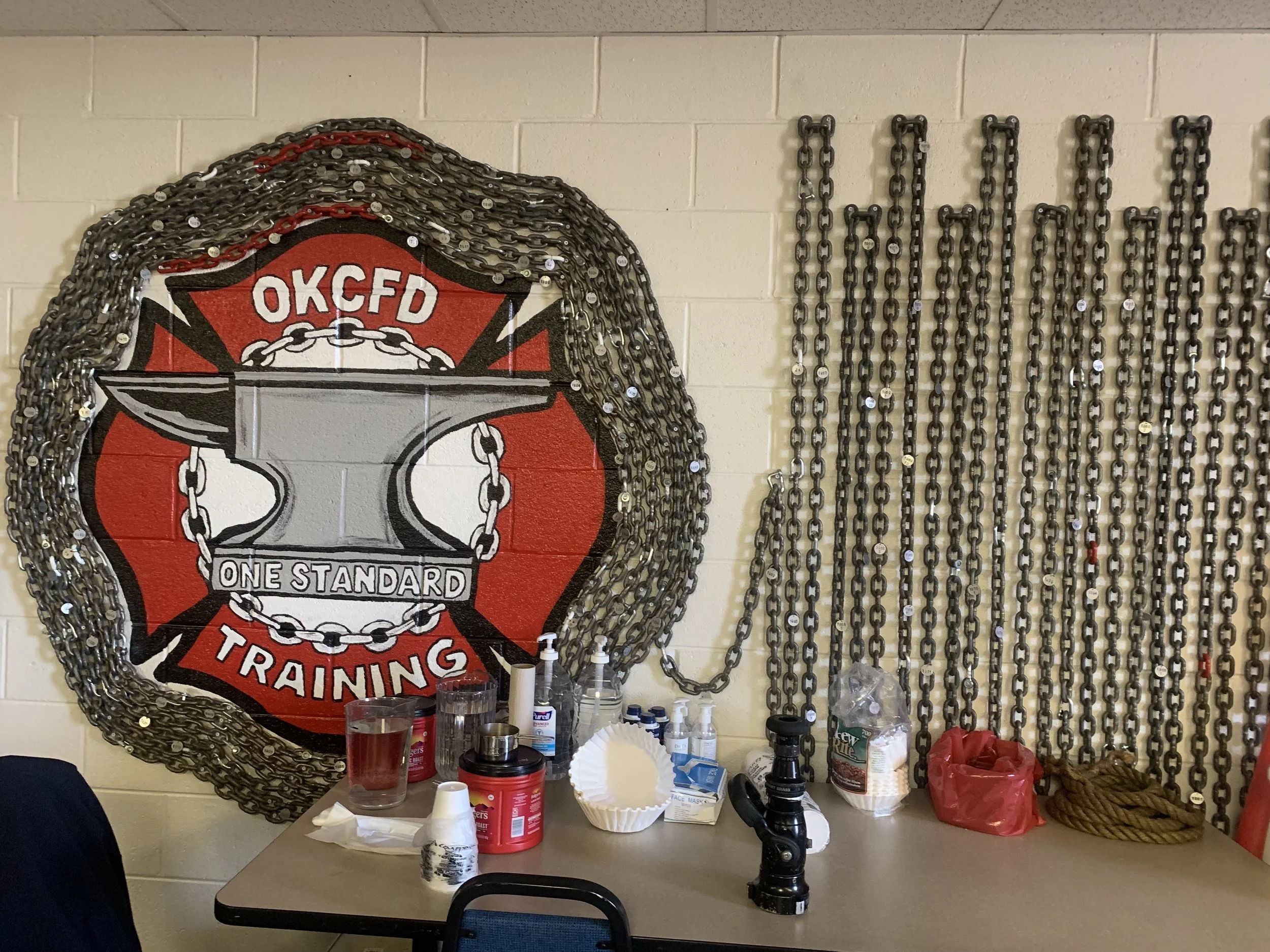 Firefighter training room with a large Oklahoma City Fire Department (OKCFD) badge on the wall, surrounded by chains and various cleaning supplies, a coffee cup, and training equipment on the table.