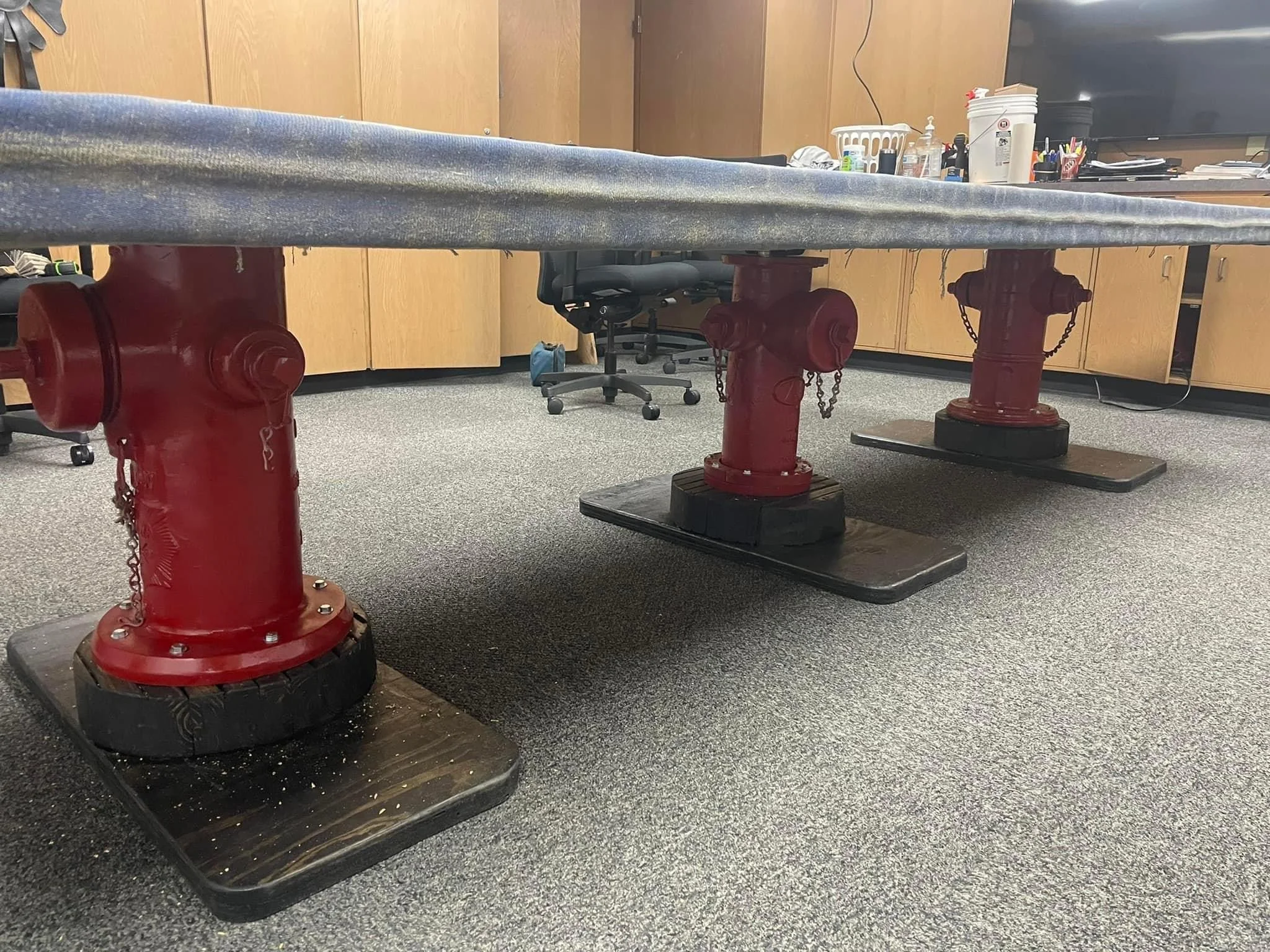 Office conference table supported by red fire hydrant bases with black wooden bases, set on a carpeted floor.