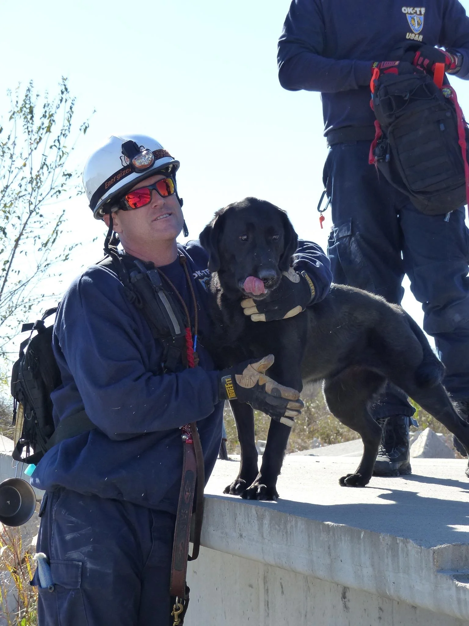 Search and rescue team member in uniform and helmet with a headlamp holding a black Labrador retriever on a concrete ledge during a search operation in daylight.