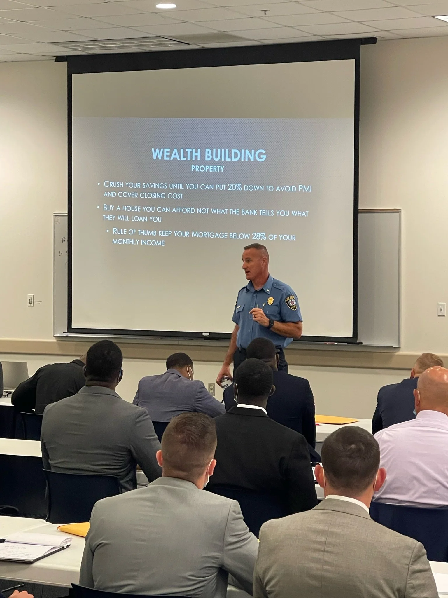 Police officer giving a presentation to a group of suited men in a conference room, with a slide titled 'Wealth Building Property' on the projection screen.