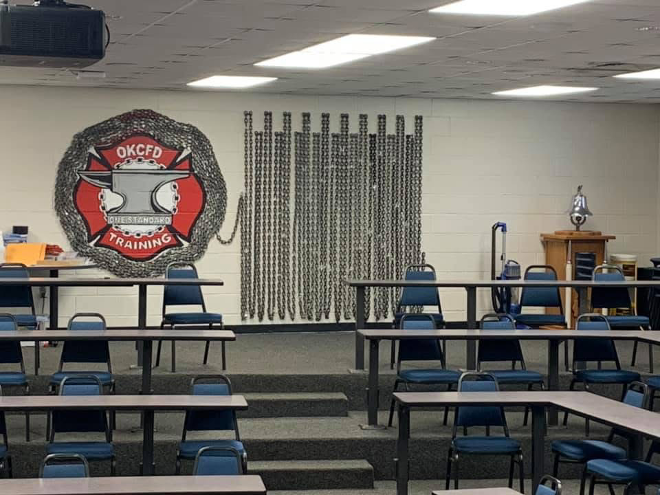 A training room with several desks and chairs, a wall display with chains, a large emblem with the text 'OKCED' and 'Training,' and a small wooden cabinet with a lamp on top.