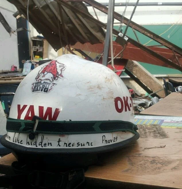 A white safety helmet with multiple stickers and handwritten notes, resting on a wooden surface in a cluttered workshop or garage.