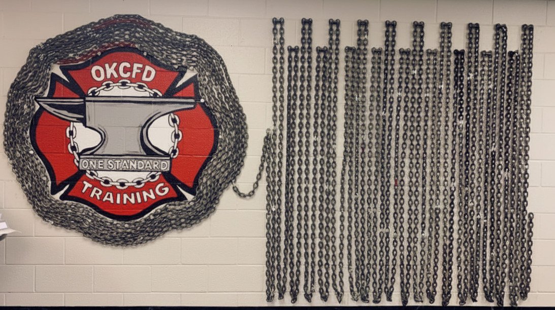 Firefighter training room with a large Oklahoma City Fire Department (OKCFD) badge on the wall, surrounded by chains and various cleaning supplies, a coffee cup, and training equipment on the table.