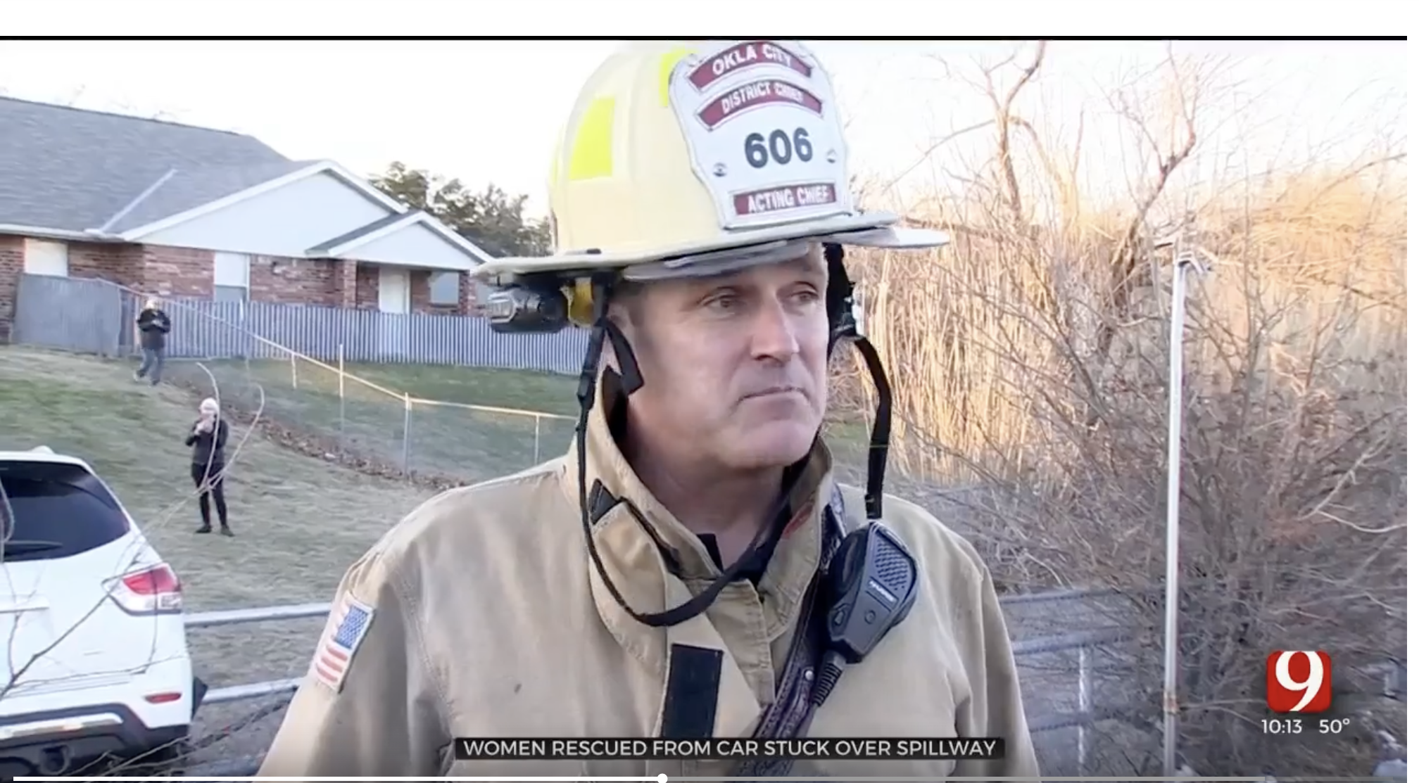A firefighter wearing a helmet and uniform stands in front of a scene that appears to involve a car accident or rescue, with a house, cars, and bystanders visible in the background, and a time and temperature display in the bottom right corner.