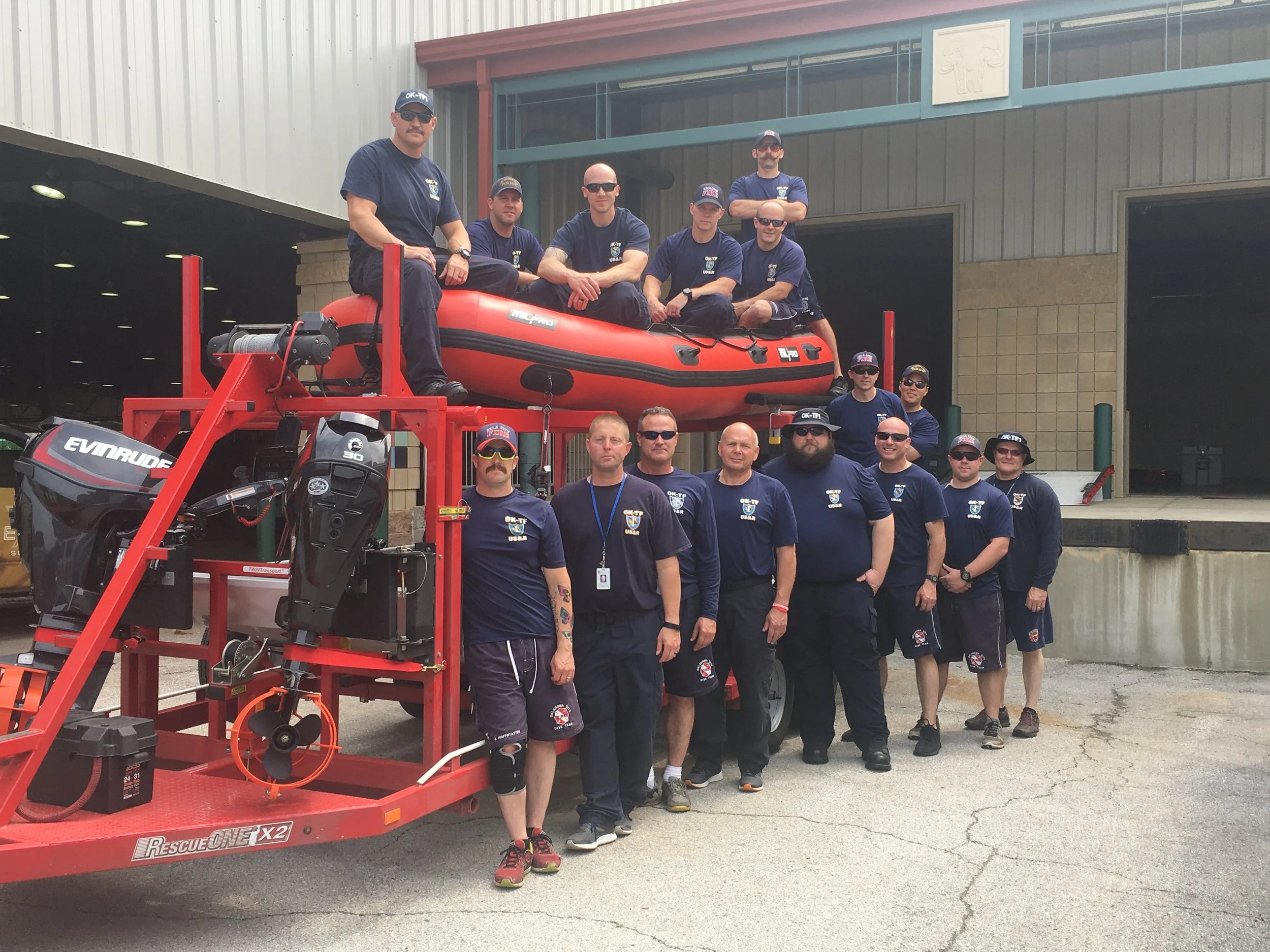 Group of emergency responders standing in front of rescue equipment, including a rescue boat and motor, outside a fire station.