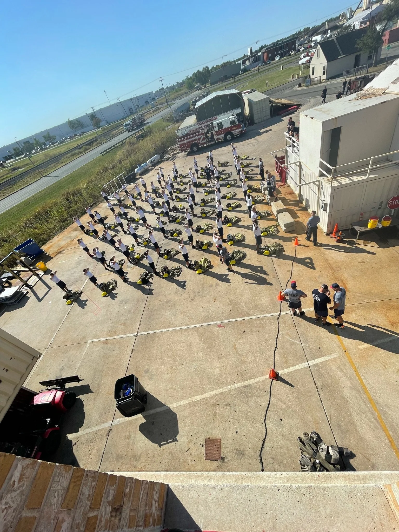 A large group of uniformed personnel practicing fire safety drills outdoors, with firefighters and equipment present, on a paved area beside a building.