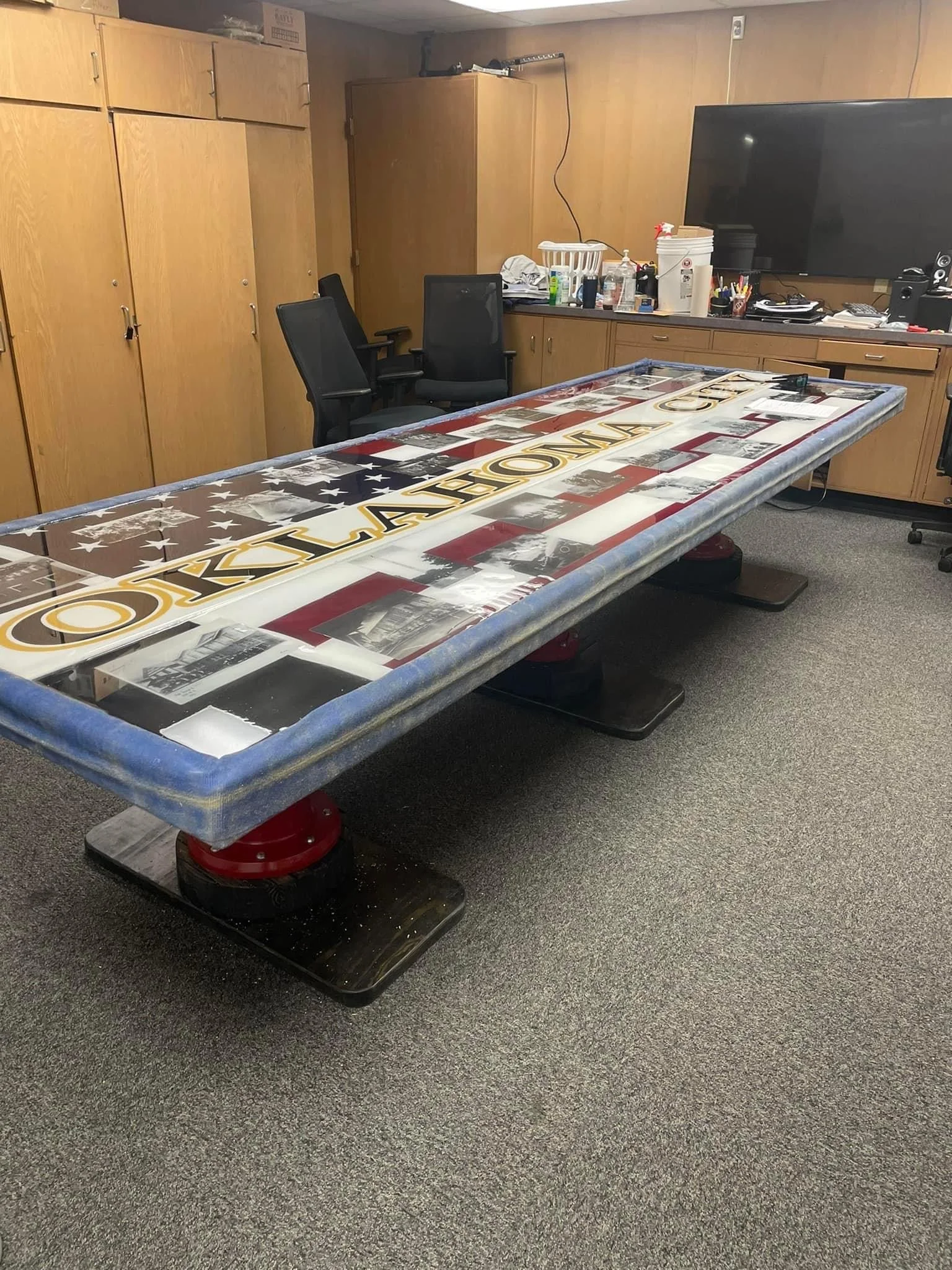 An office room with a large tabletop covered with a glass display, featuring a patriotic American flag design and text, surrounded by chairs and office supplies, with wooden cabinets and a large TV on the wall.