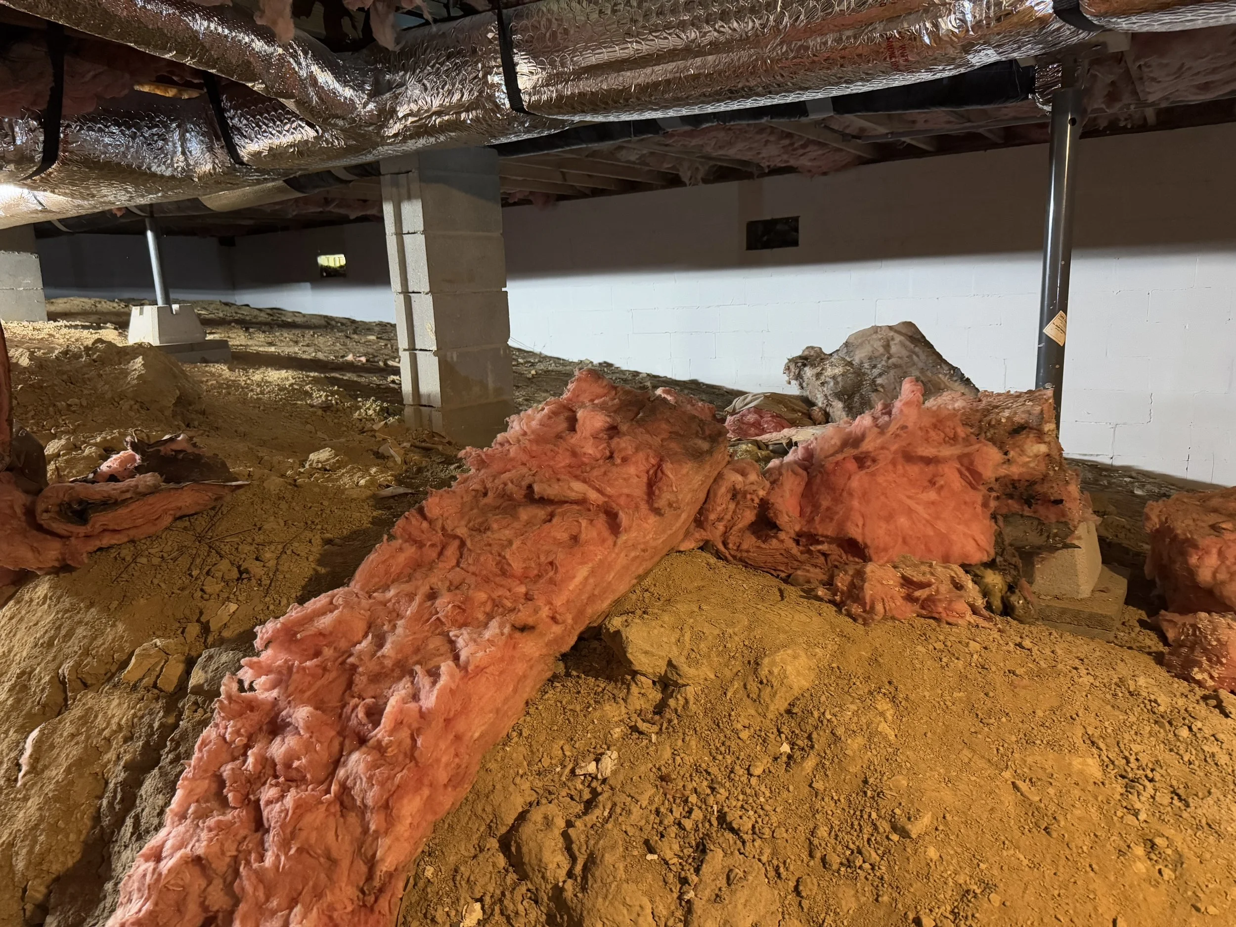 Construction site with exposed insulation and dirt, unfinished basement or crawl space, with support beams and concrete blocks.