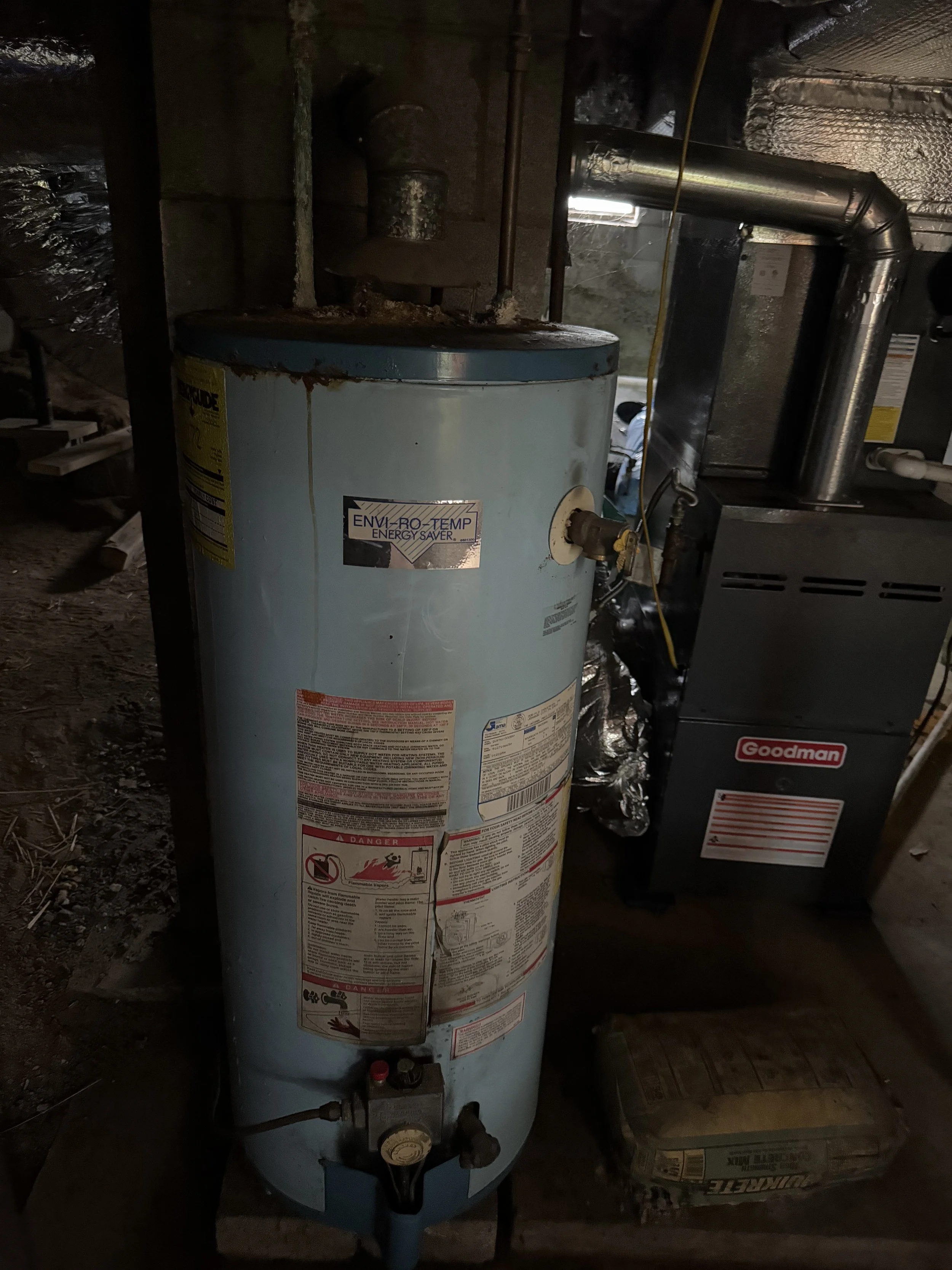 A blue water heater tank with warning labels, connected to pipes in a basement, with a Goodman furnace nearby.