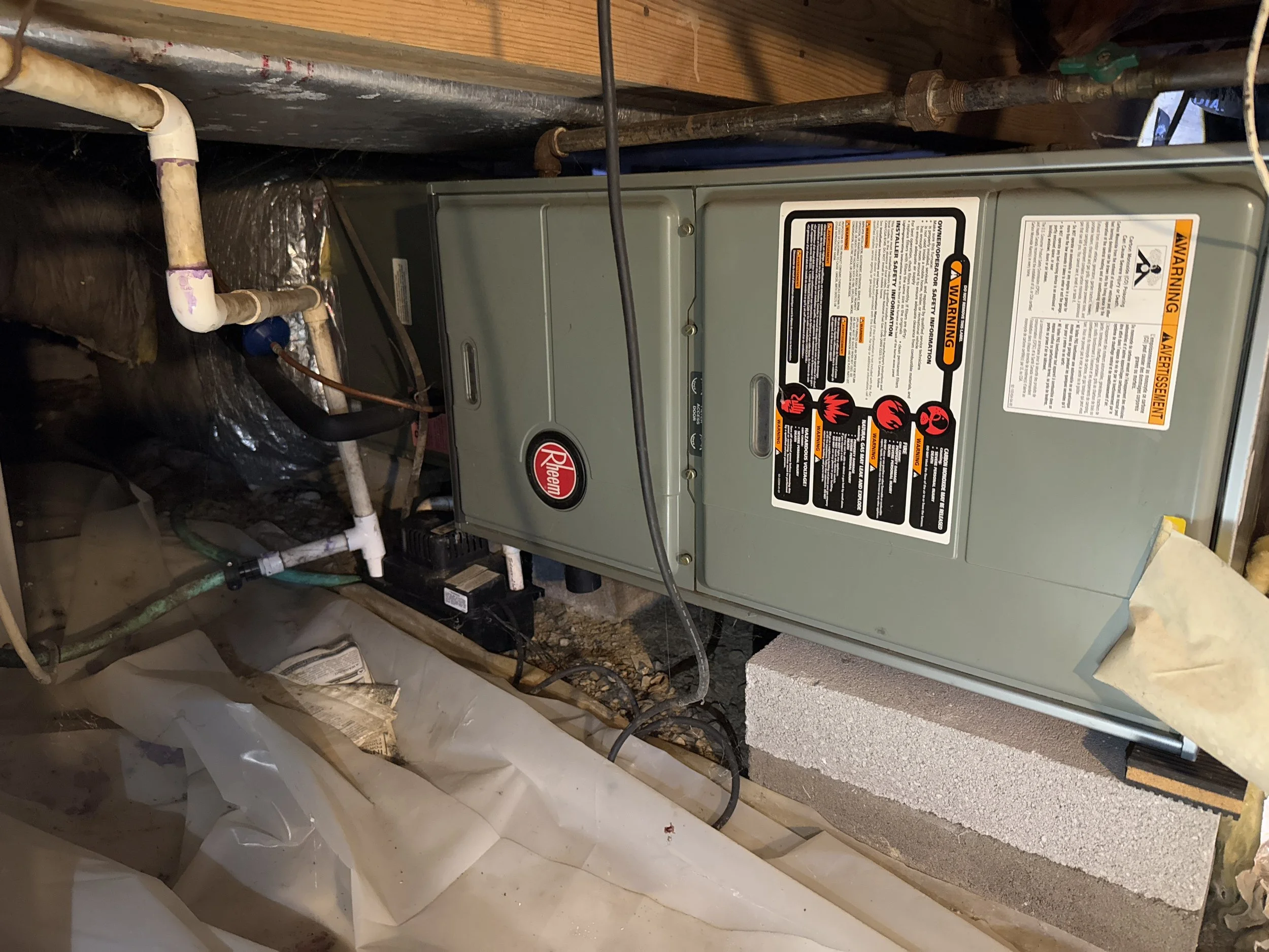 A Rheem furnace unit installed in a basement, with piping and wiring connected around it, mounted on a concrete block with insulation material nearby.