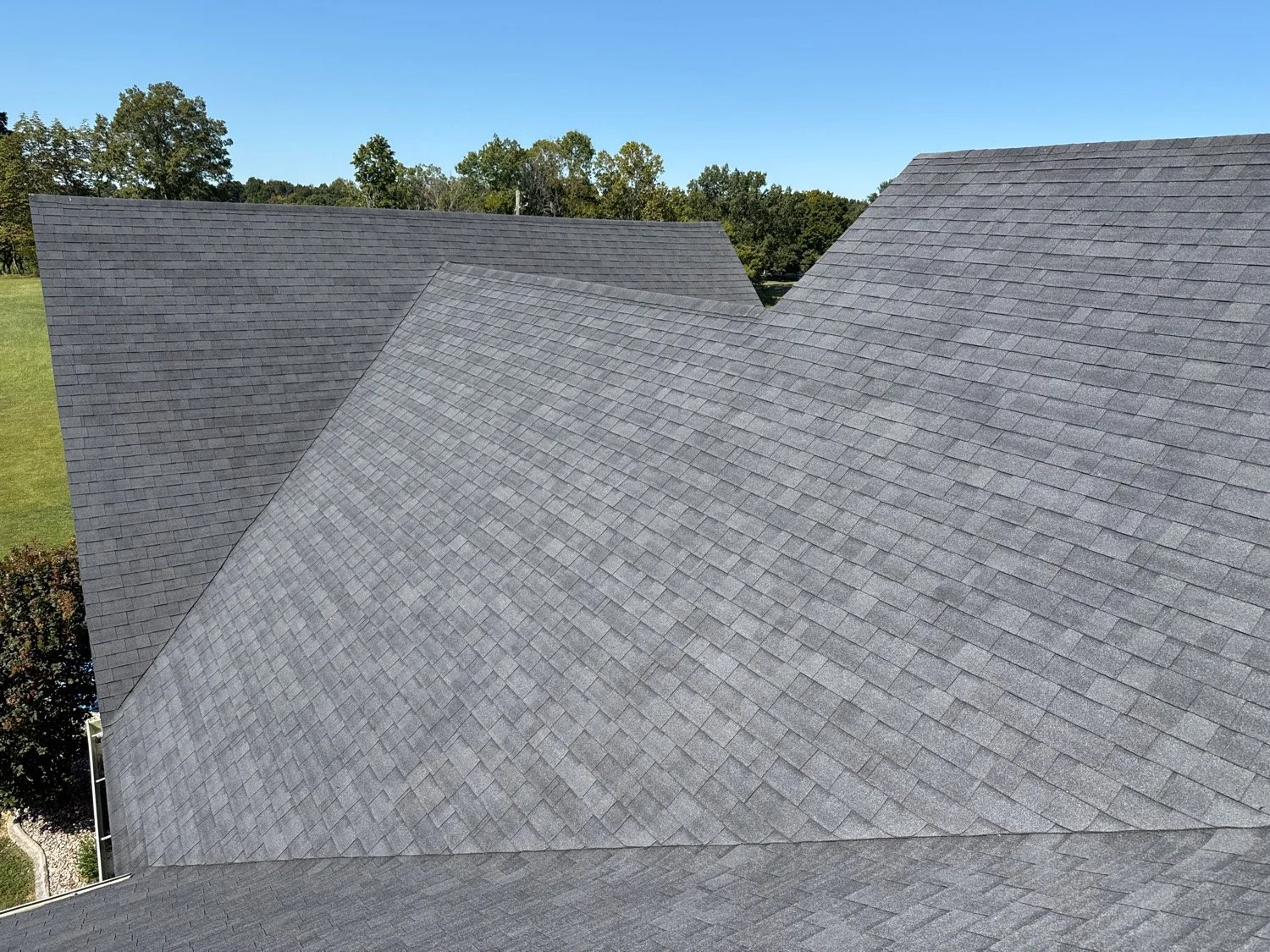 Upward view of multiple gray shingle roofs with trees and blue sky in the background.