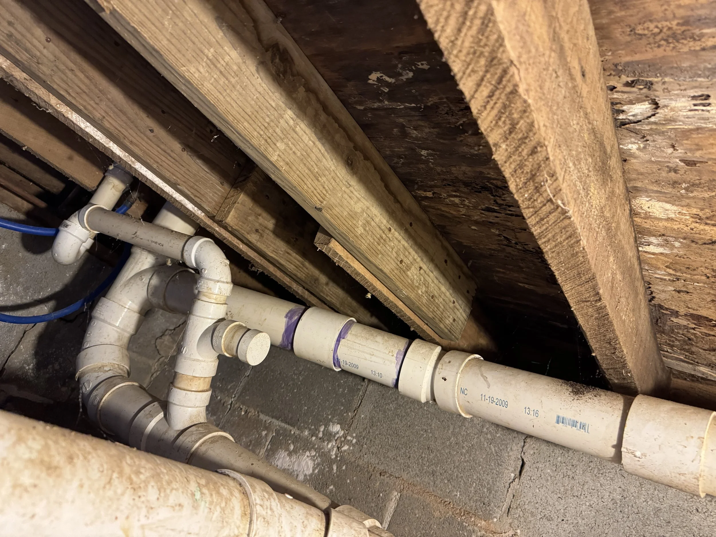 Under construction area showing exposed plumbing pipes, concrete blocks, and overhanging wooden beams.