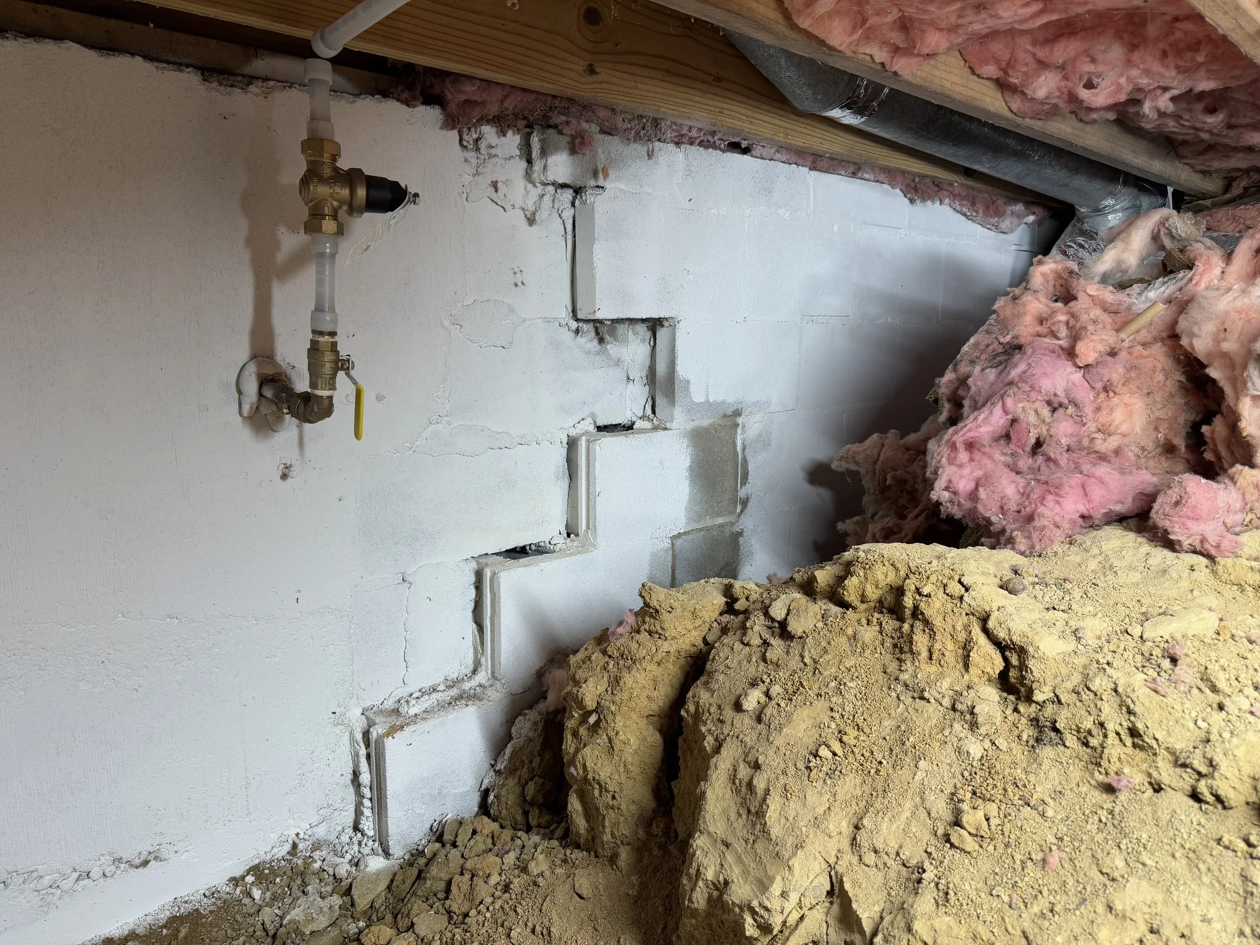 Construction site with exposed wall with pipe fitting, pink insulation, and pile of dirt and yellow gravel.