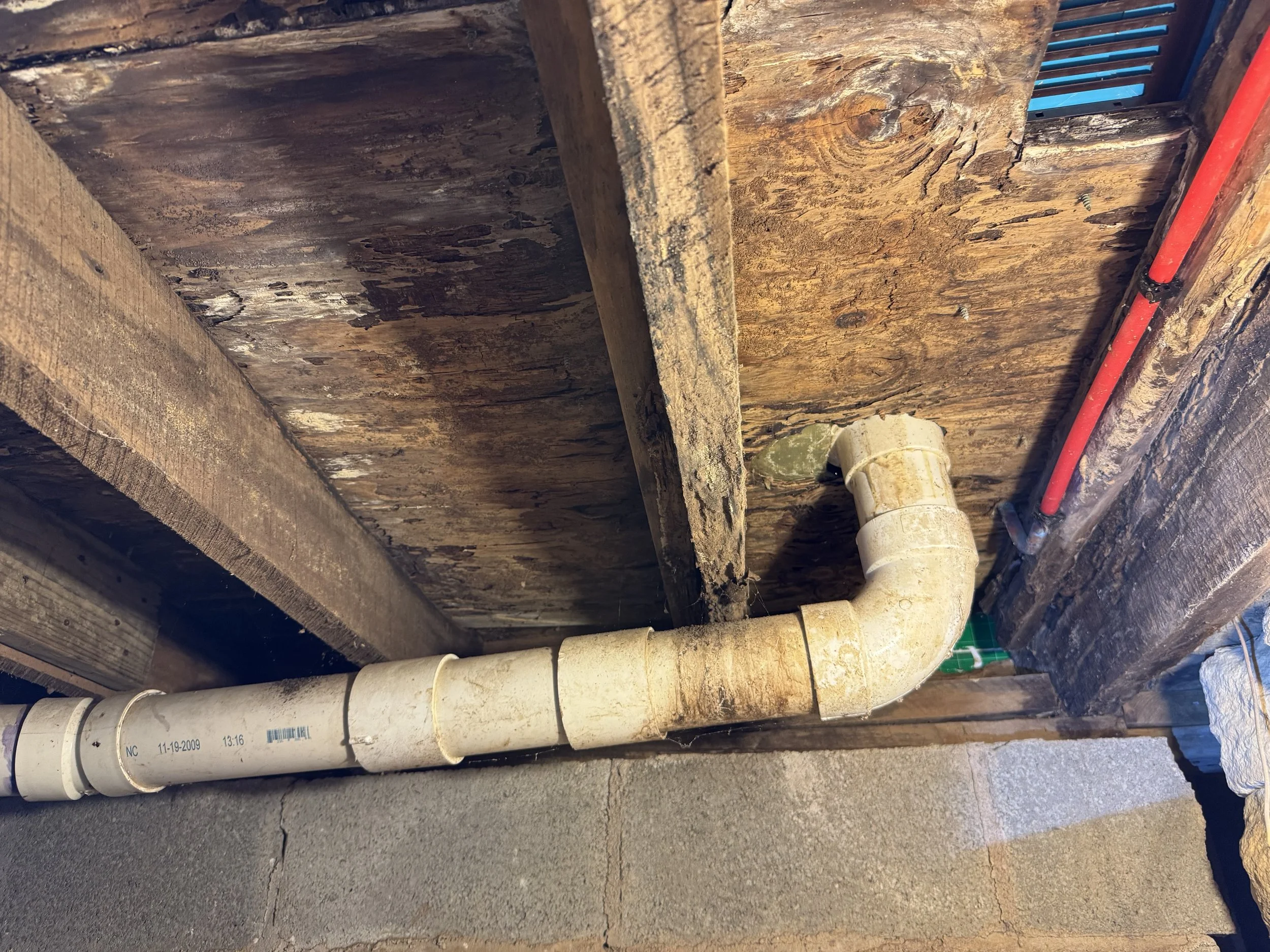 Close-up of exposed plumbing pipes in a construction area with wooden framing and walls. The pipes appear dirty and used, with some dirt and stains visible.