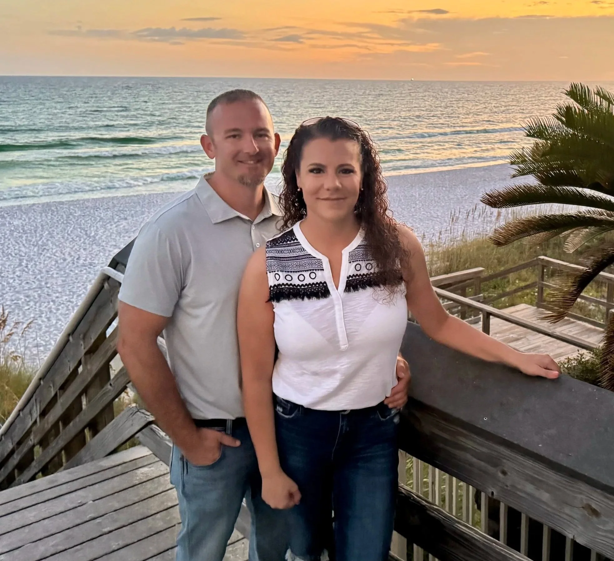 A man and woman standing on a wooden deck with a beach and ocean at sunset in the background.
