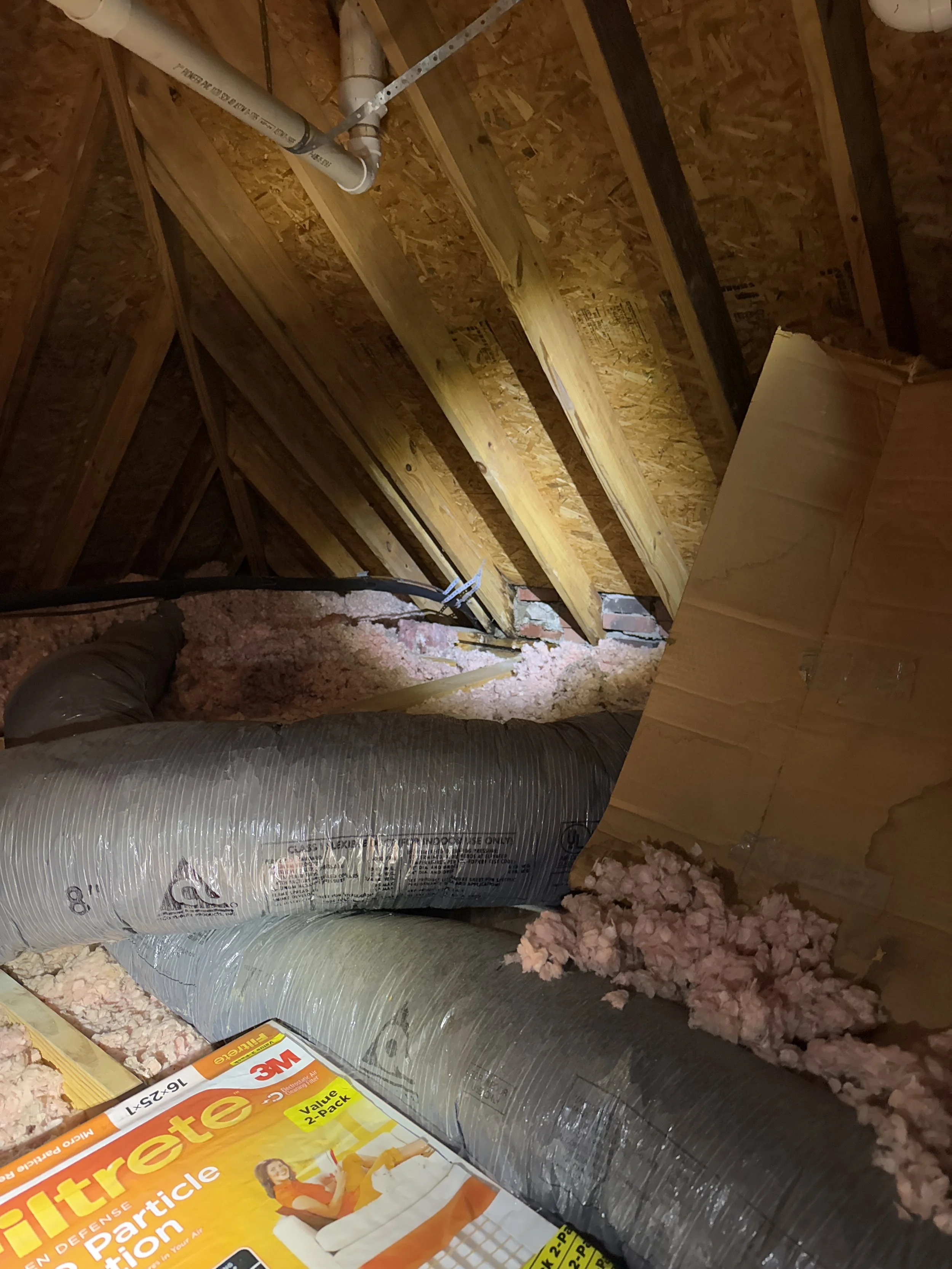 Interior of an attic with exposed insulation, a large duct, wooden beams, and some cardboard. A section of brick wall is visible in the background.
