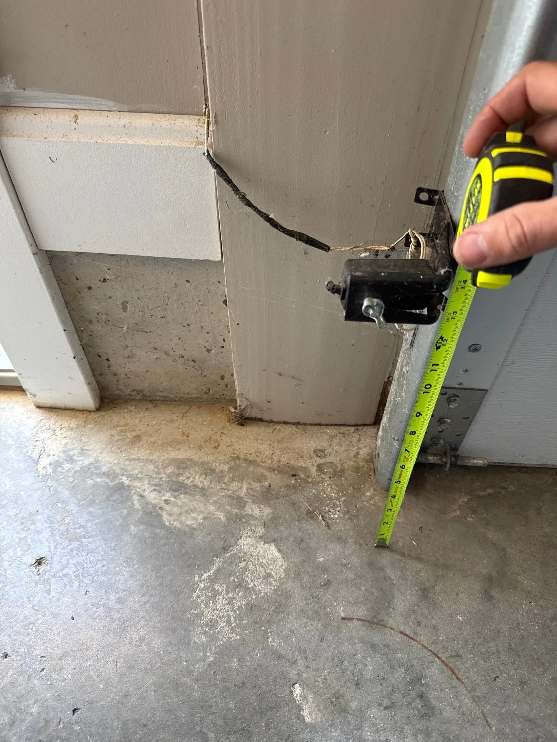 Close-up of a person measuring the height of a garage door opener motor with a tape measure. The motor is mounted at the top of a garage wall near the ceiling, with wires and mounting brackets visible. The garage floor shows dirt and dust.