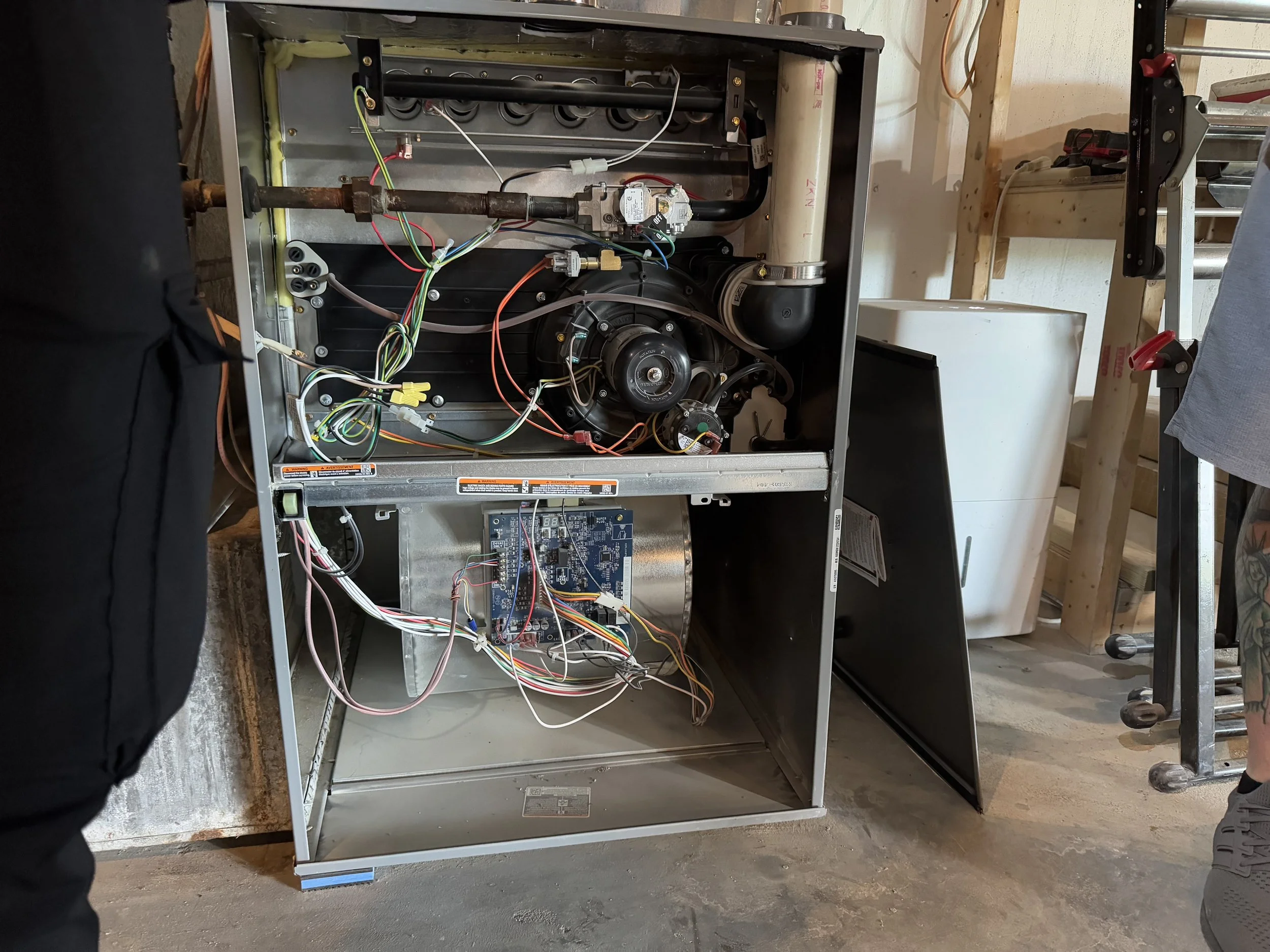 The front panel of an open washing machine with exposed internal components, including wiring, motor, and control circuit board, situated in a workshop or garage setting.