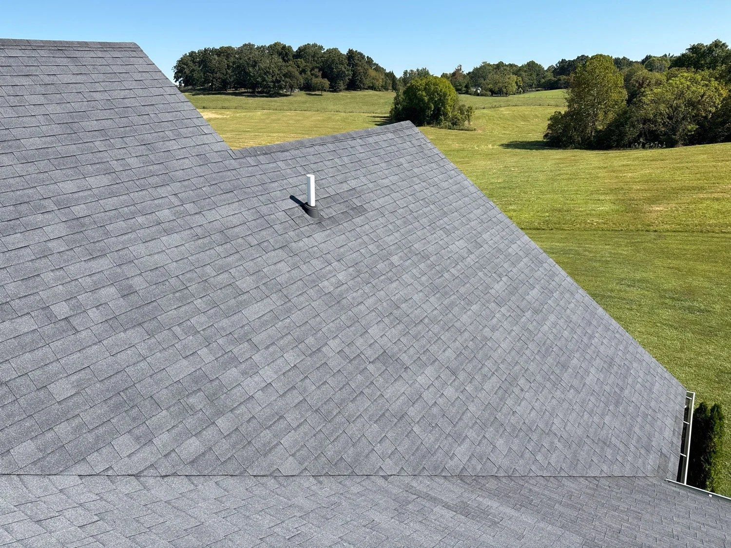 A bird's eye view of a gray shingled roof with a vent pipe, surrounded by green fields and trees under a clear blue sky.