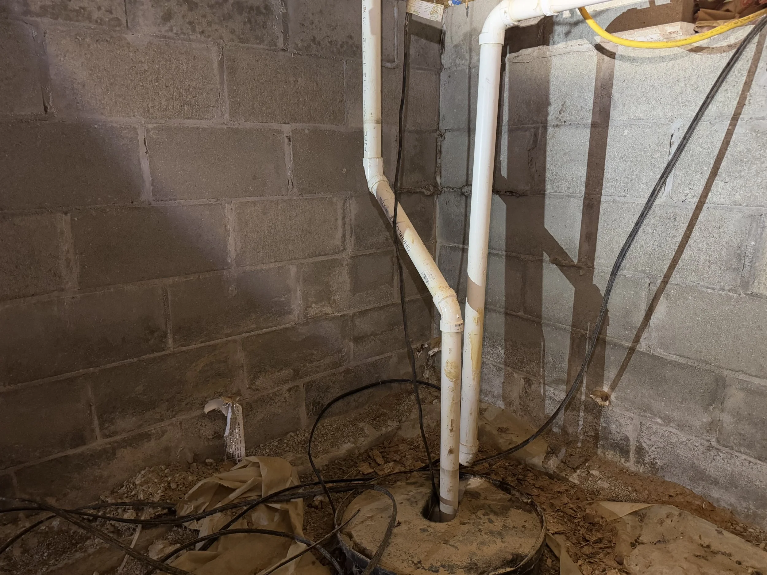 PVC pipes and black electrical wires in a basement or utility room with unfinished concrete walls and dirt floor.