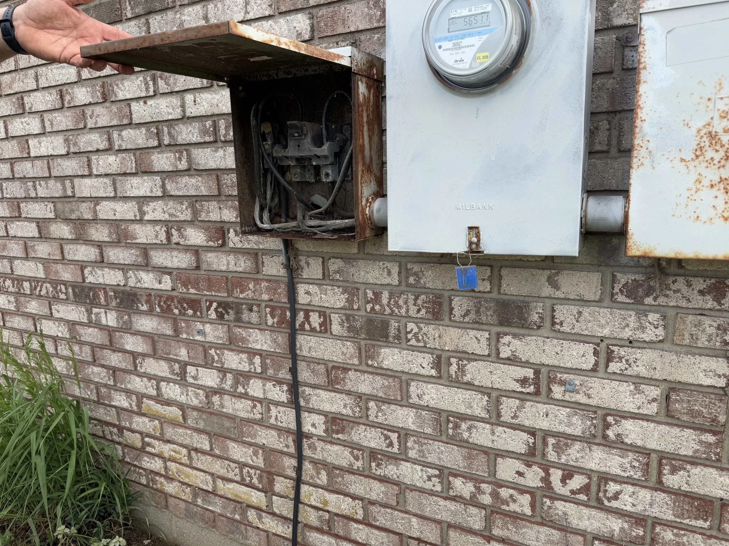 A brick wall with a rusty open electrical box, a meter box, and a rusty streetlight control box. An electrical wire hangs down from the open box.