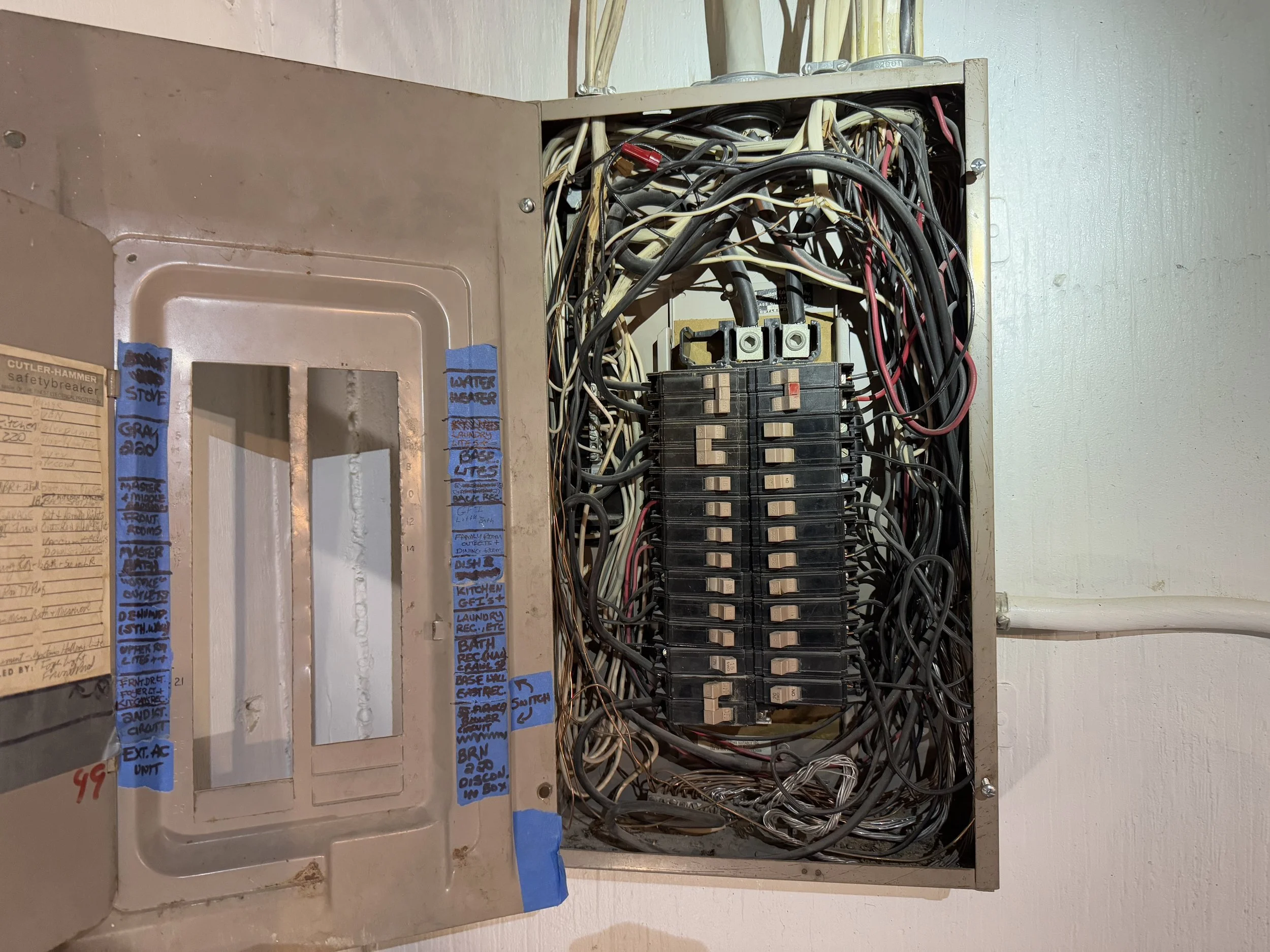 Open electrical breaker box with wires and circuit breakers inside, with handwritten labels on the inside of the door for different circuits.