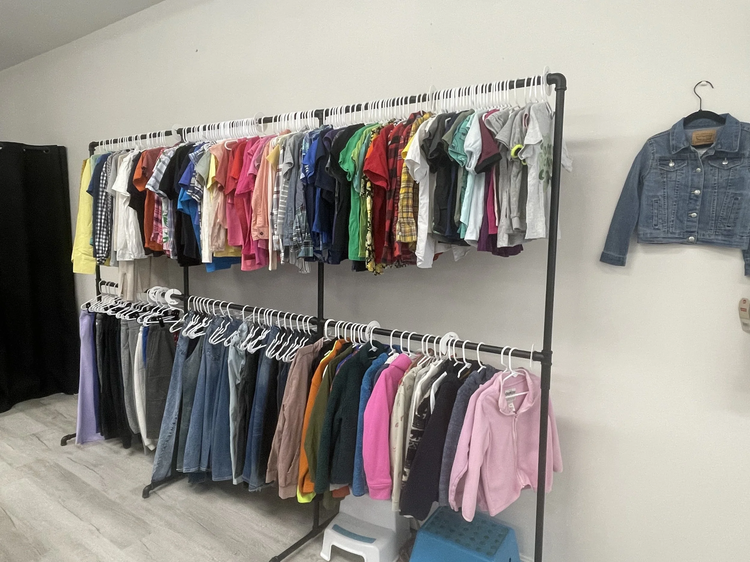 Clothing rack with colorful shirts on top and various pants and jackets below, in a room with a white wall and wooden floor.