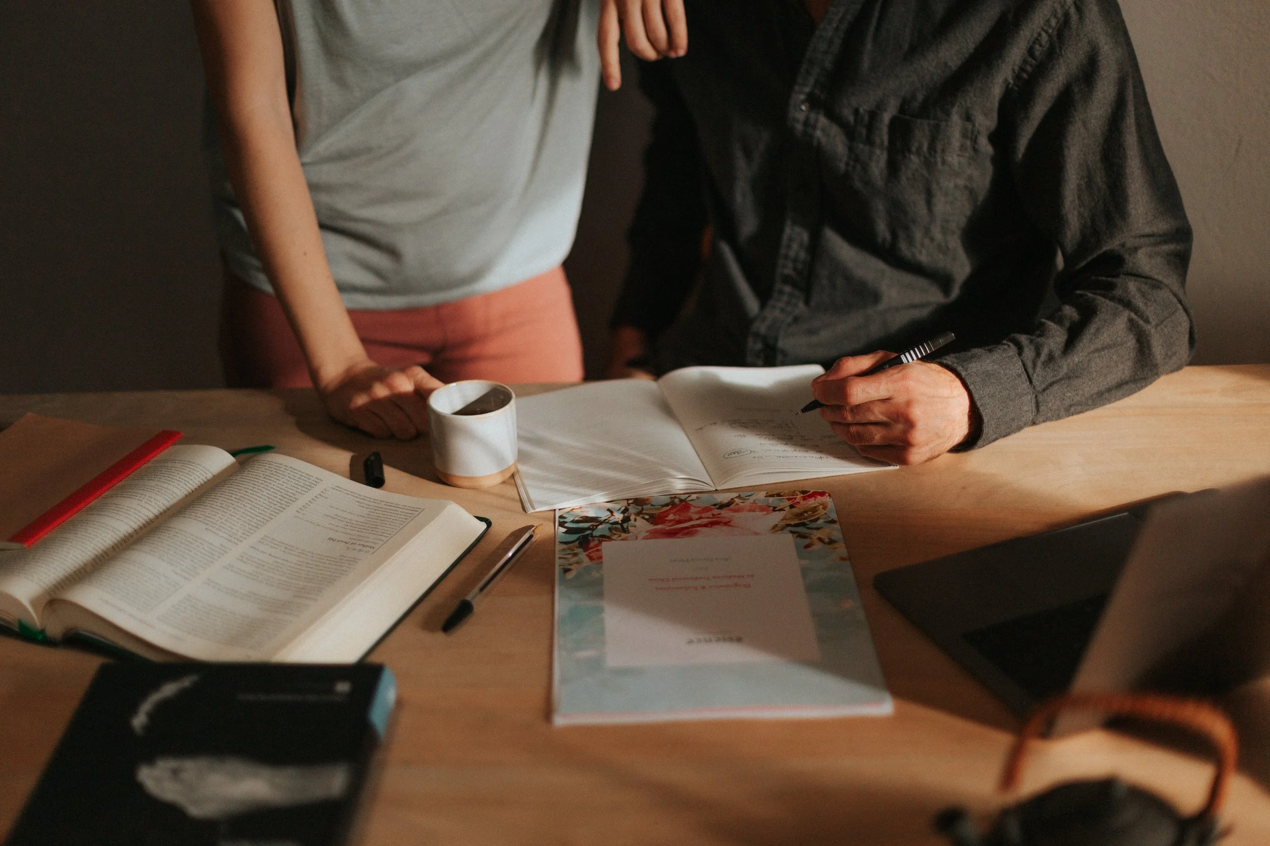Personas revisando documentos y tomando notas en una mesa de trabajo con libros, cuaderno, taza y laptop.