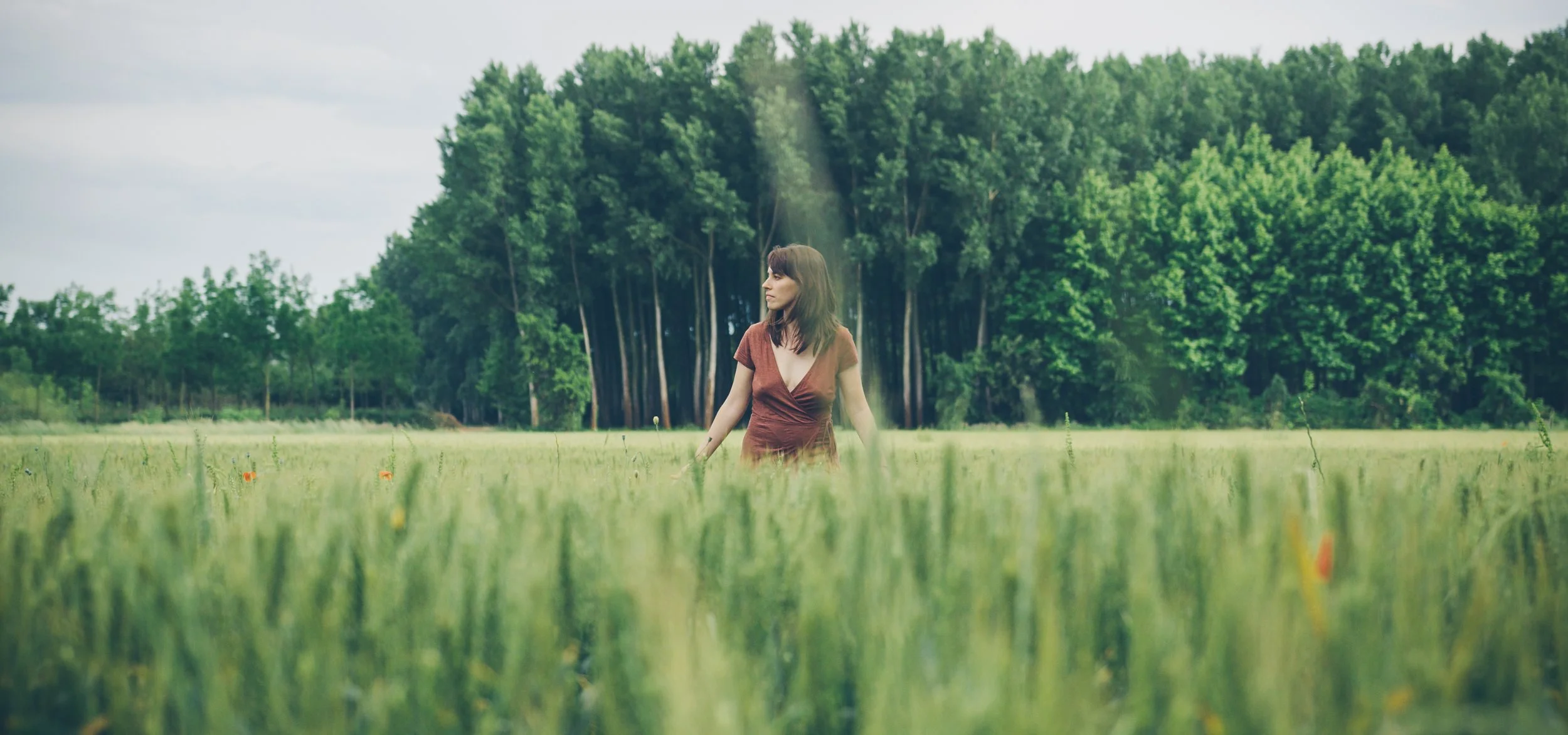 Mujer de cabello largo y vestido marrón en un campo de hierba con árboles de fondo