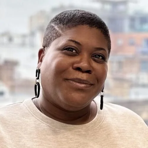 Close-up of Simone Payne - Powell. Chartered MCIPD, MCMI, Fractional HR Director,  a woman with short hair, wearing earrings, smiling with an urban cityscape in the background.