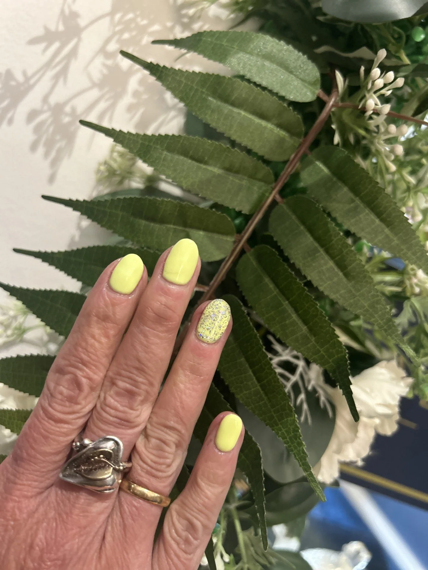 A person's left hand with yellow painted nails and a glitter accent nail resting on green leaves and white flowers.