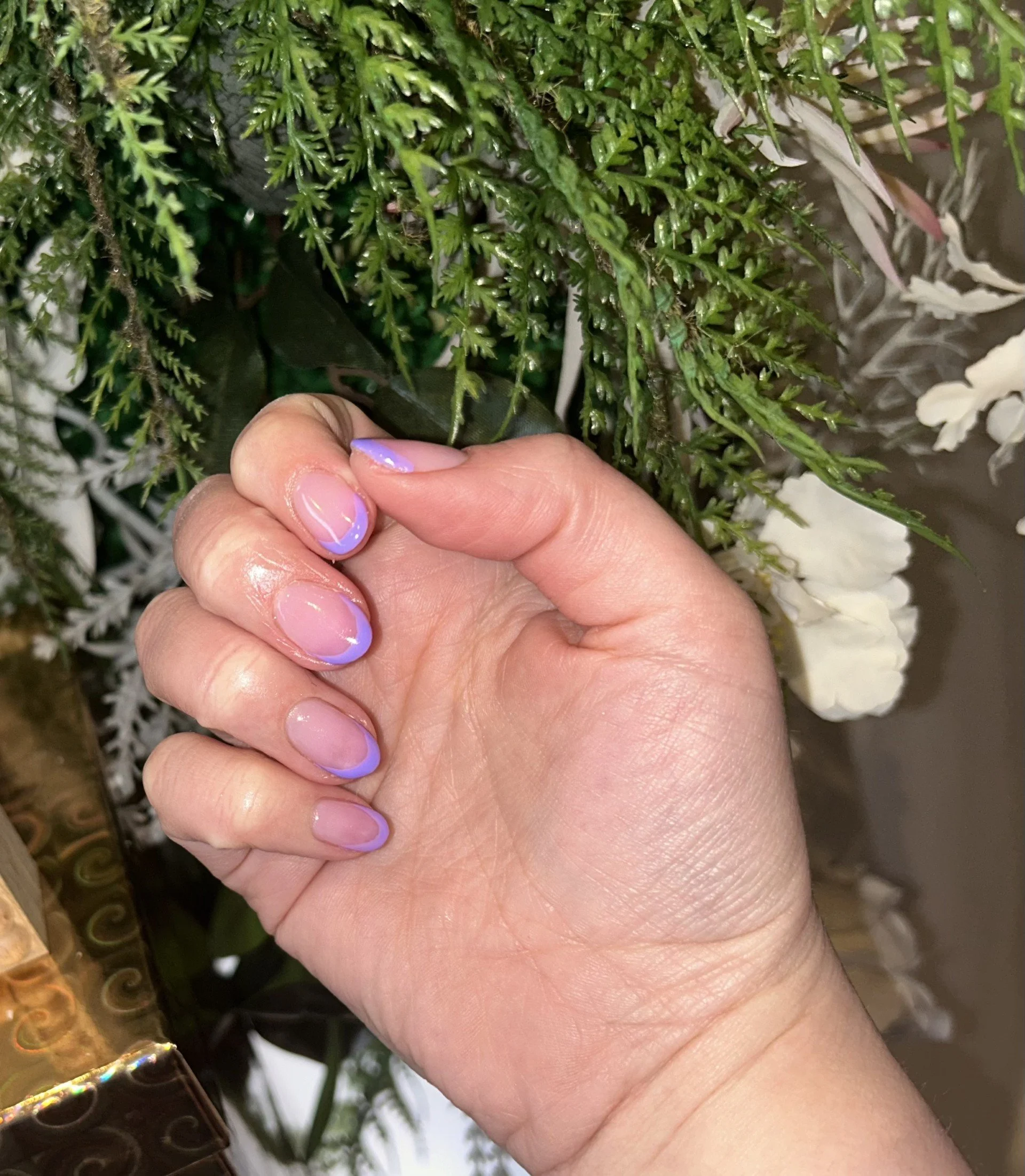 Close-up of a hand with manicured nails featuring a lavender and clear polish design, positioned against a background of green foliage and white flowers.