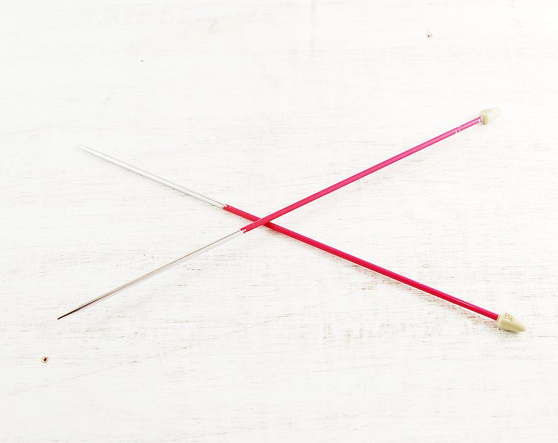 Two sewing pins with pink and silver shafts crossing on a white wooden surface.