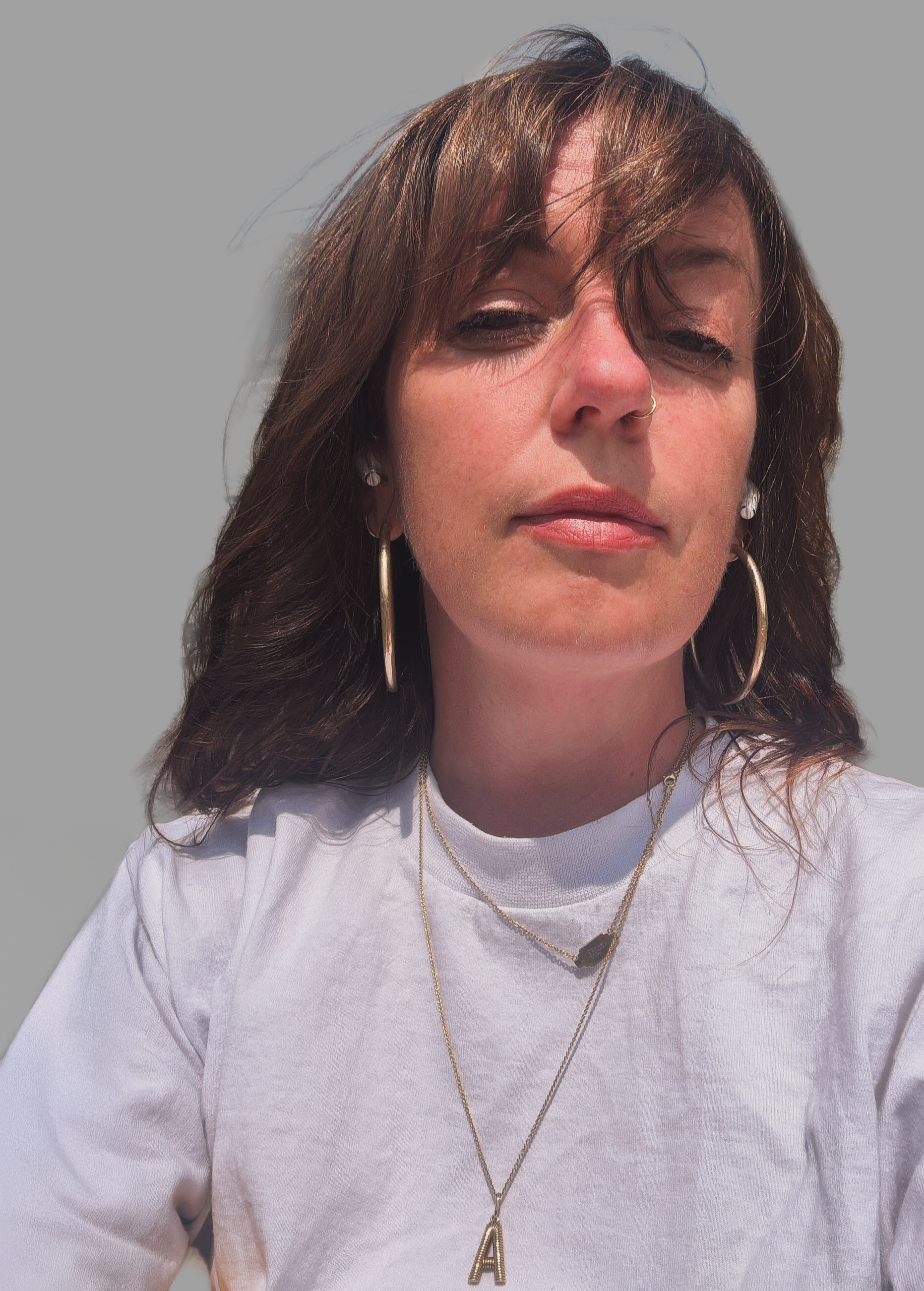 Close-up of a woman with brown hair wearing hoop earrings, layered necklaces, and a white shirt, standing against a plain gray background.