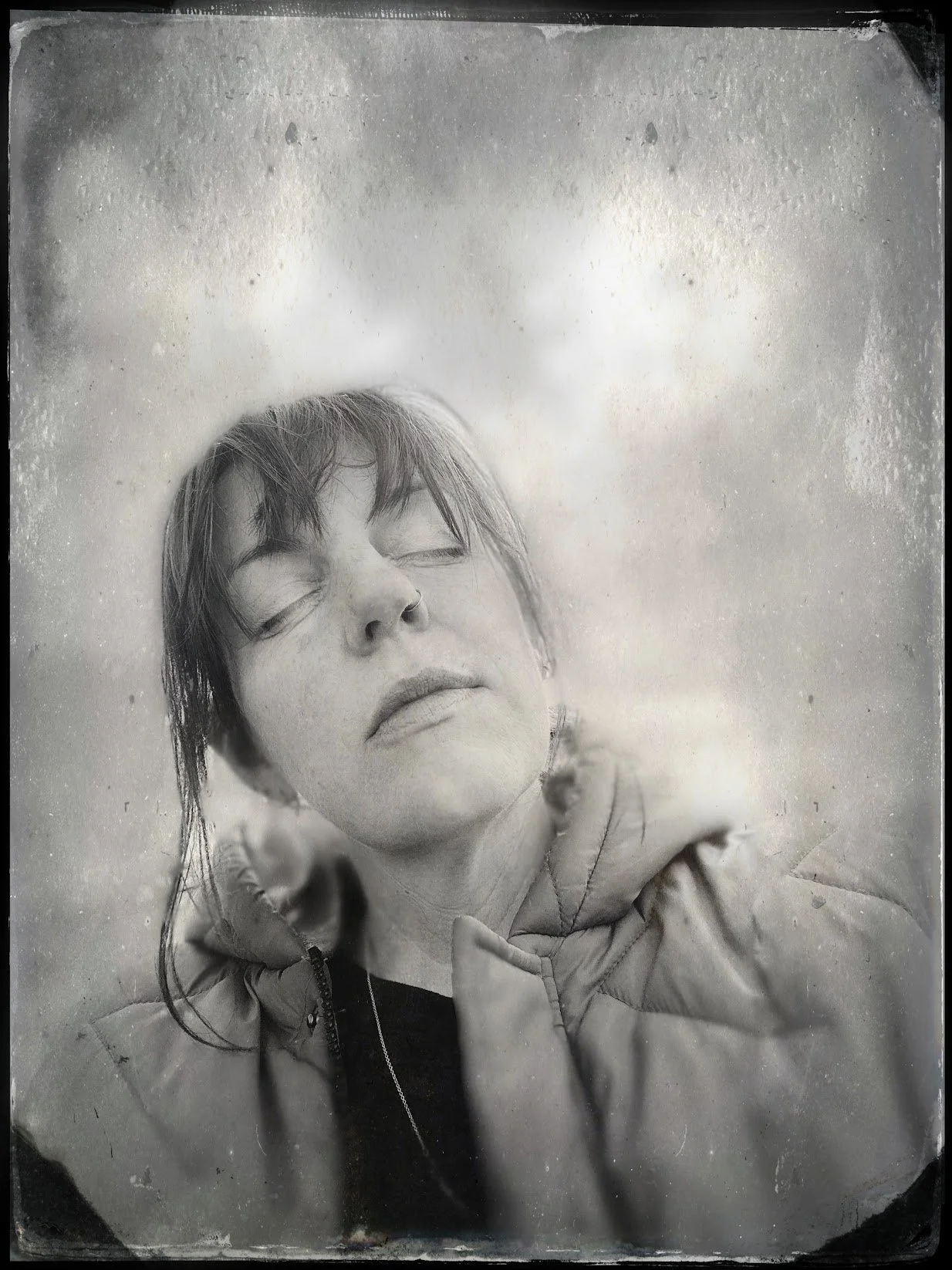 Black and white photo of a woman with short hair and freckles, eyes closed, looking contemplative, wearing a hoodie and jacket, with a second person partially visible behind her.