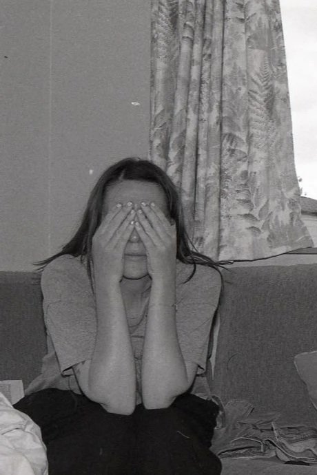 A woman sitting on a couch with her hands covering her face, in front of a patterned curtain.