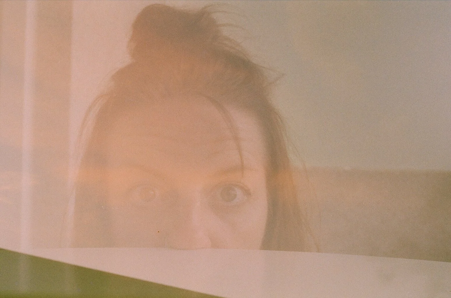 Photograph of a woman's face, with her hair tied up in a bun, reflected on a glass surface created by overlapping layers.