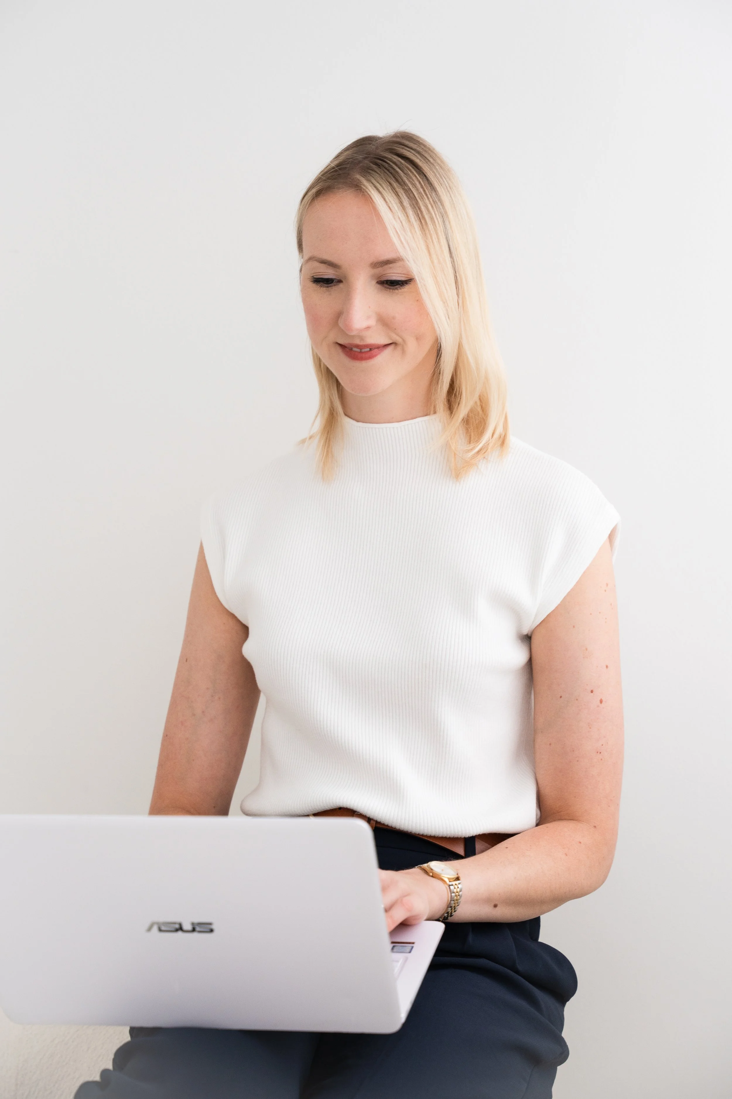 Eine Frau mit blonden Haaren sitzt an einem weißen Schreibtisch und arbeitet an einem weißen Asus-Laptop, trägt ein weißes Oberteil und eine goldene Armbanduhr.