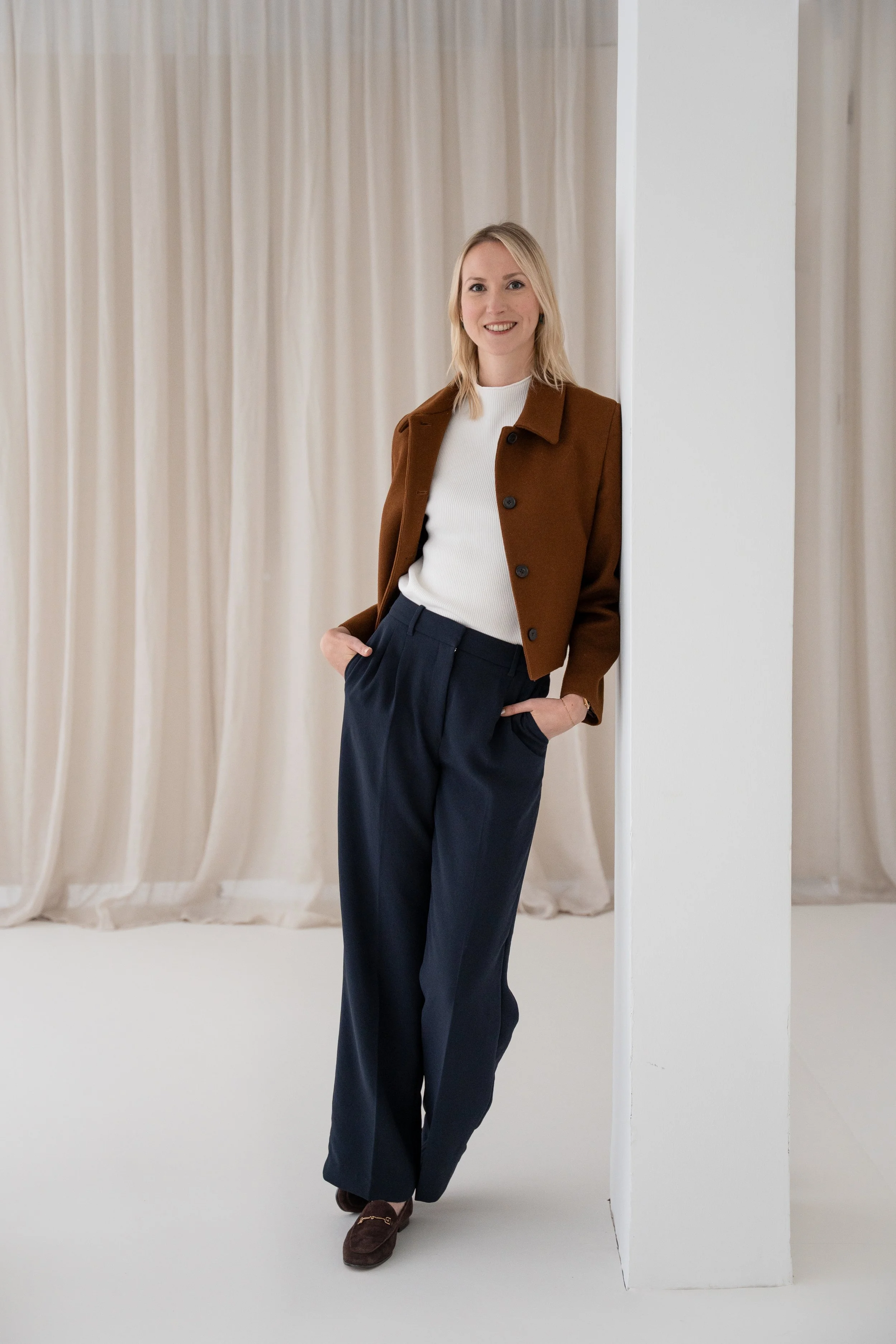 Junge Frau in Business-Kleidung lehnt an einer weißen Wand, lächelt, trägt braune Jacke, weiße Bluse, dunkle Hose und braune Schuhe vor einem beige Curtain-Hintergrund.