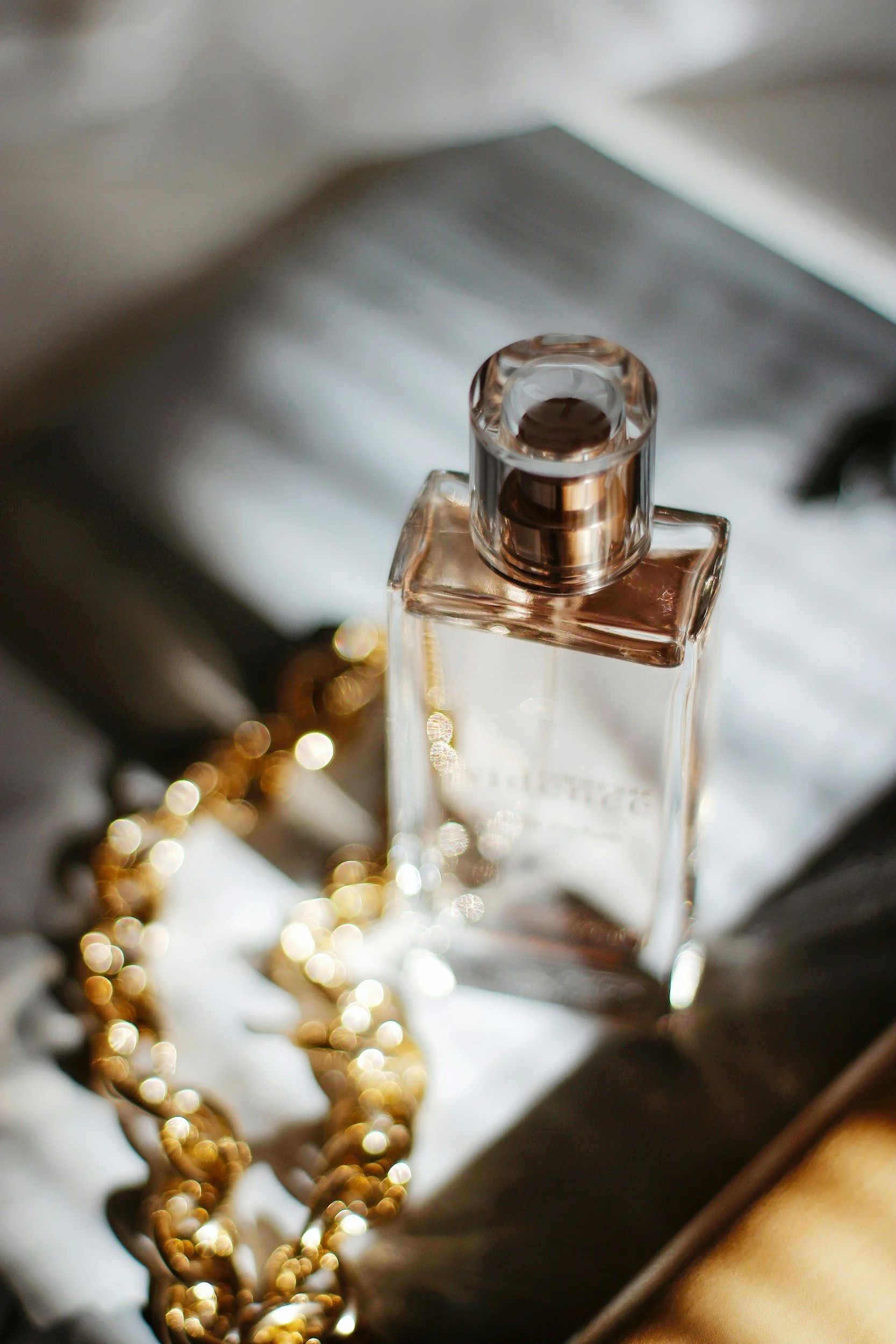 Parfum rose dans un flacon en verre, accompagnée d'un collier en chaîne dorée, posés sur un livre avec lumière naturelle.