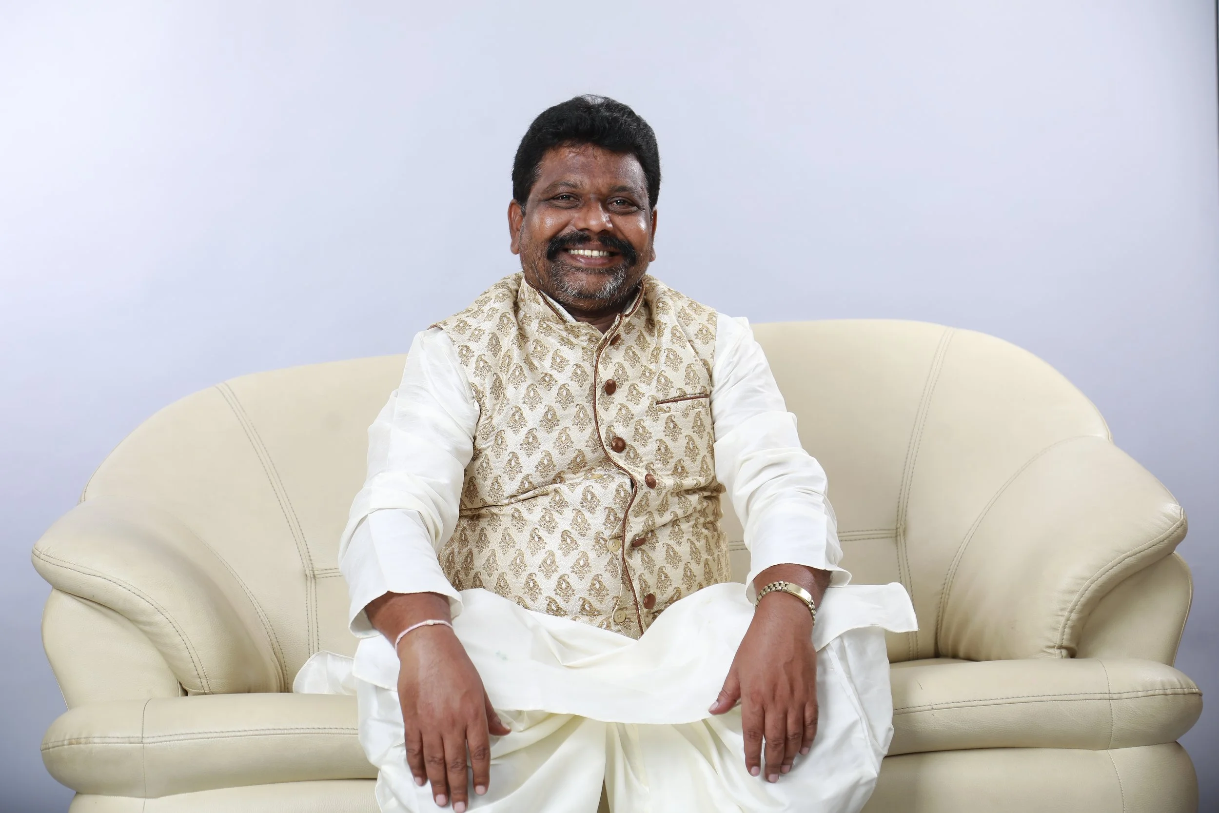 A smiling man with a beard, wearing traditional Indian attire, sitting on a cream-colored sofa against a plain white background.