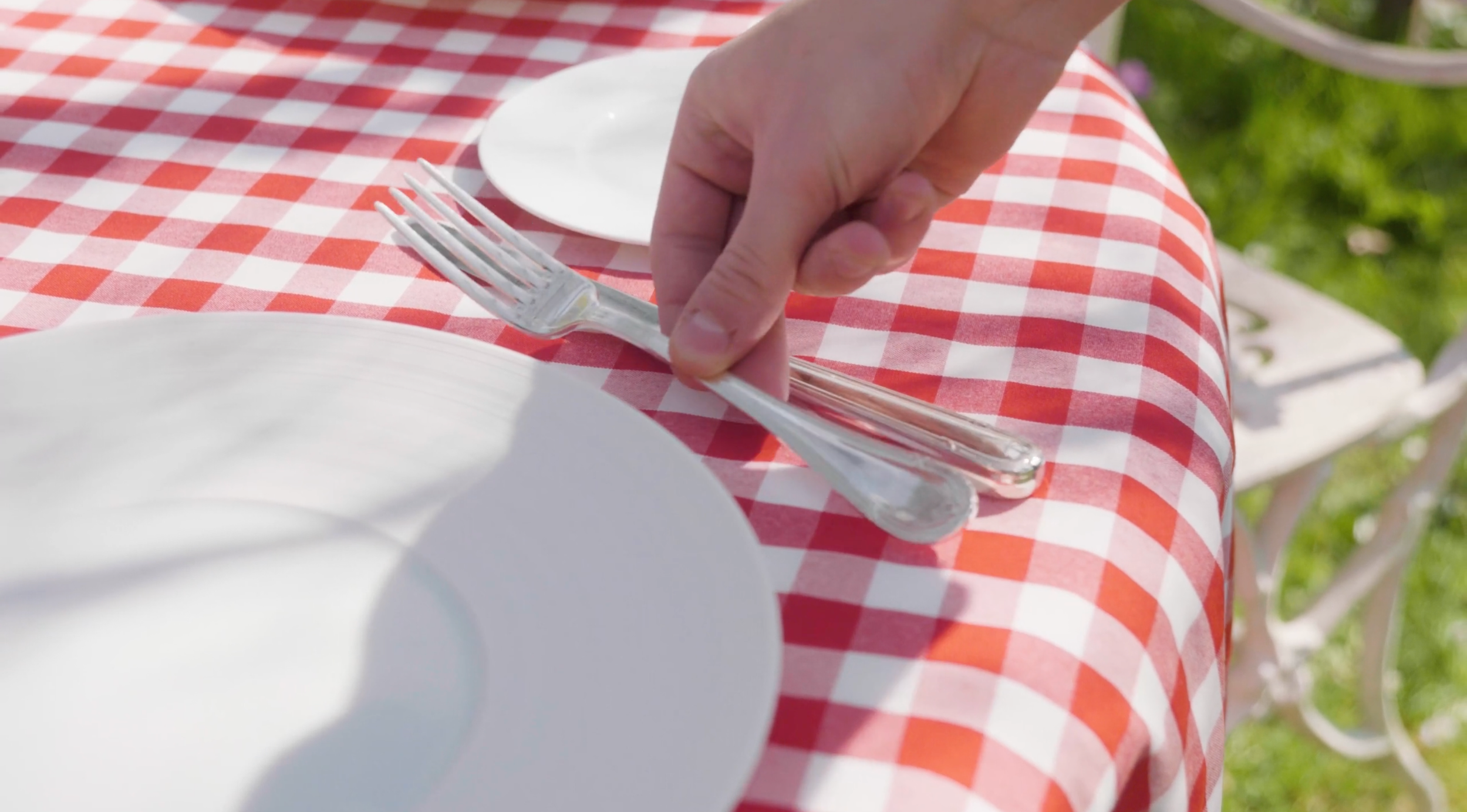 Une main pose une fourchette sur une nappe à carreaux rouges et blancs, avec des assiettes blanches sur la table.