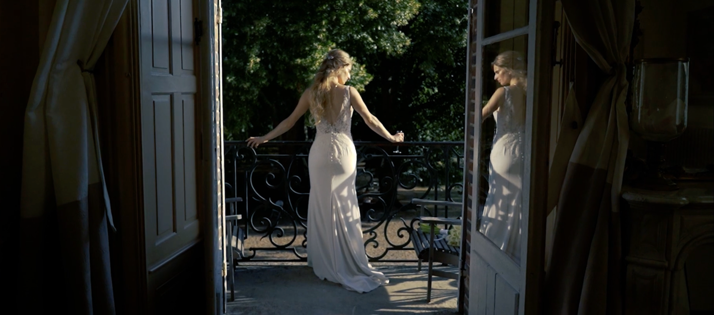 Une femme en robe de mariage blanche se tient sur un balcon, regardant à l'extérieur, avec une reflection dans la porte en verre à côté d'elle.